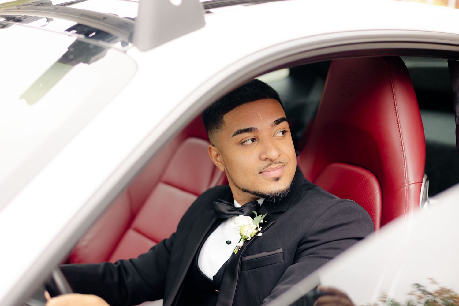 Formal portrait of person in tuxedo seated in luxury car with red leather interior.