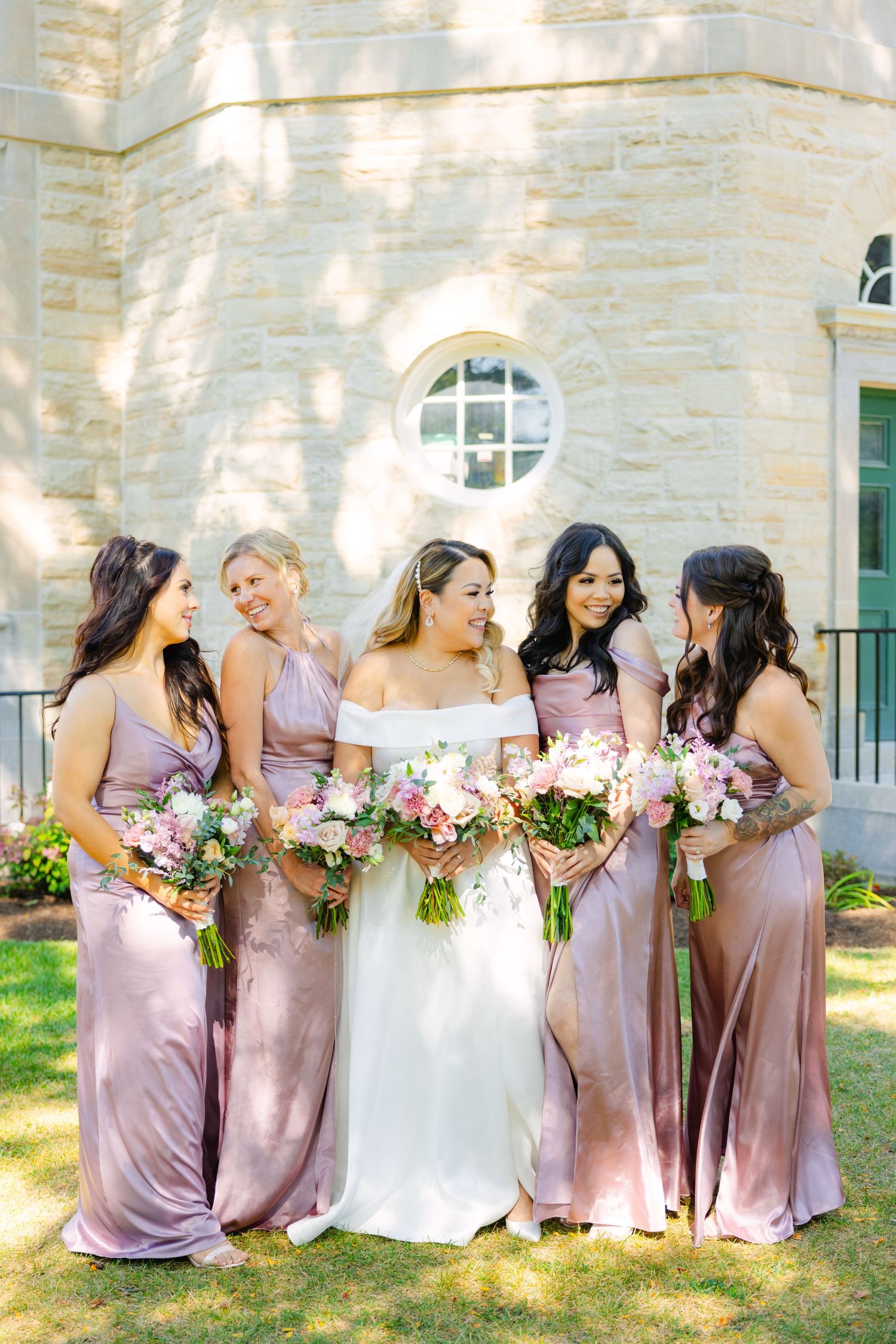 Bride and bridesmaids in pink gowns hold floral bouquets against stone church exterior.