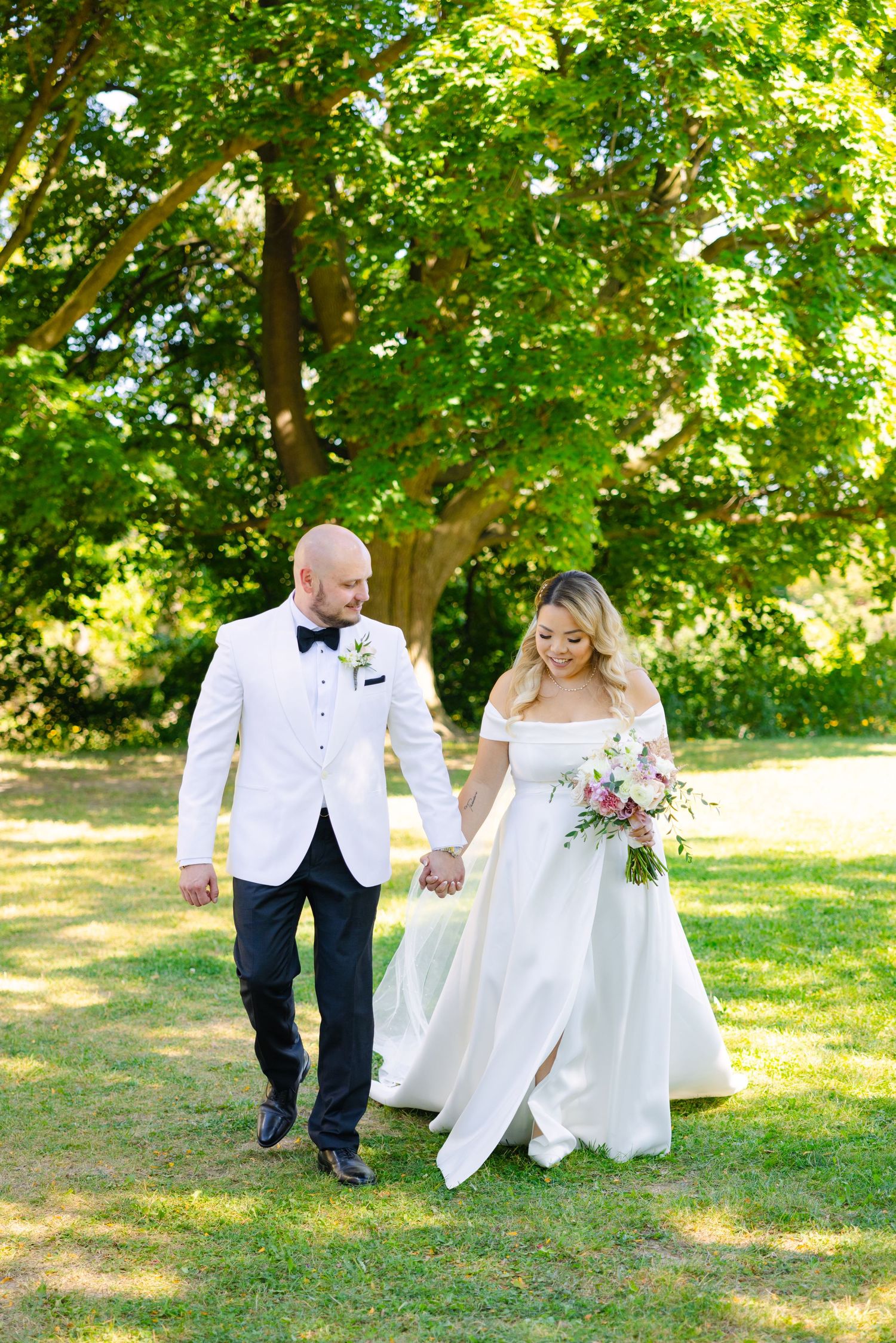 Newlyweds stroll together on grass path under canopy of green leaves.