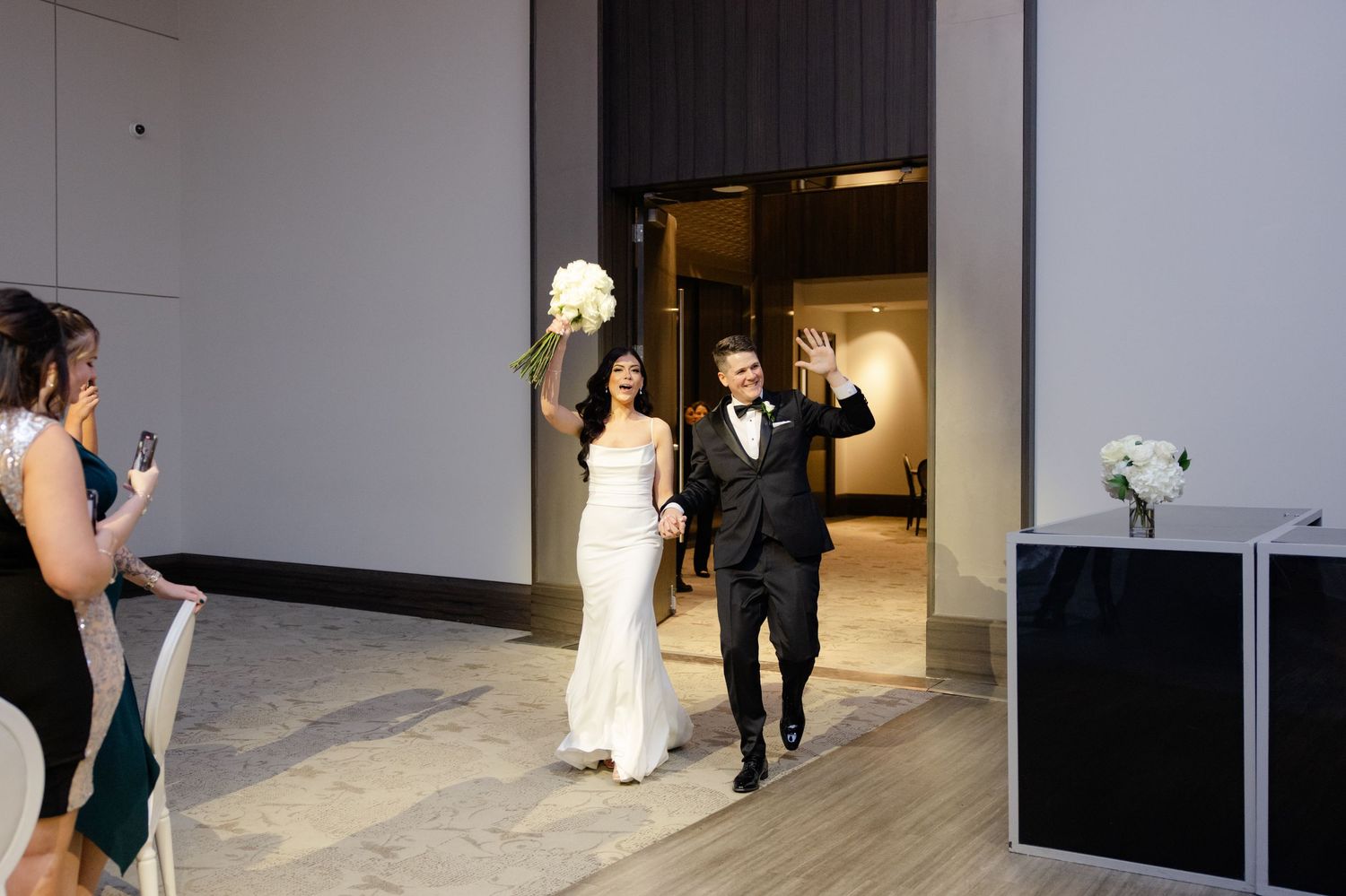 Newlyweds make their celebratory entrance through modern venue doorway with guests watching.