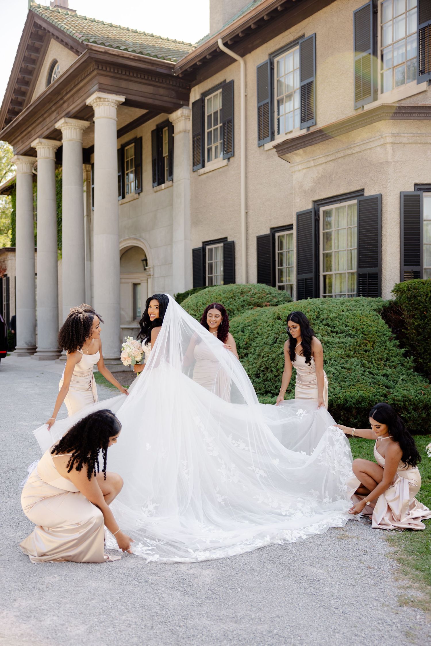 Bridesmaids arrange long cathedral veil outside historic building with columns.