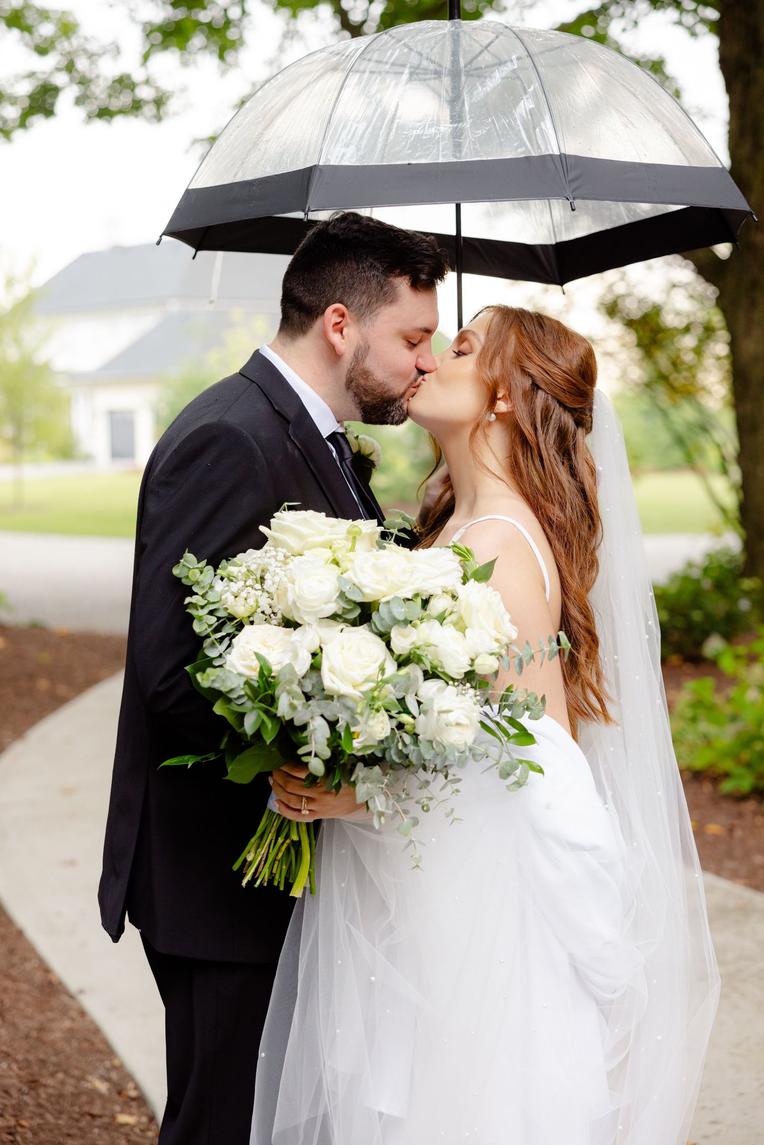 A couple kisses under a clear umbrella during their rainy day wedding ceremony with white roses.