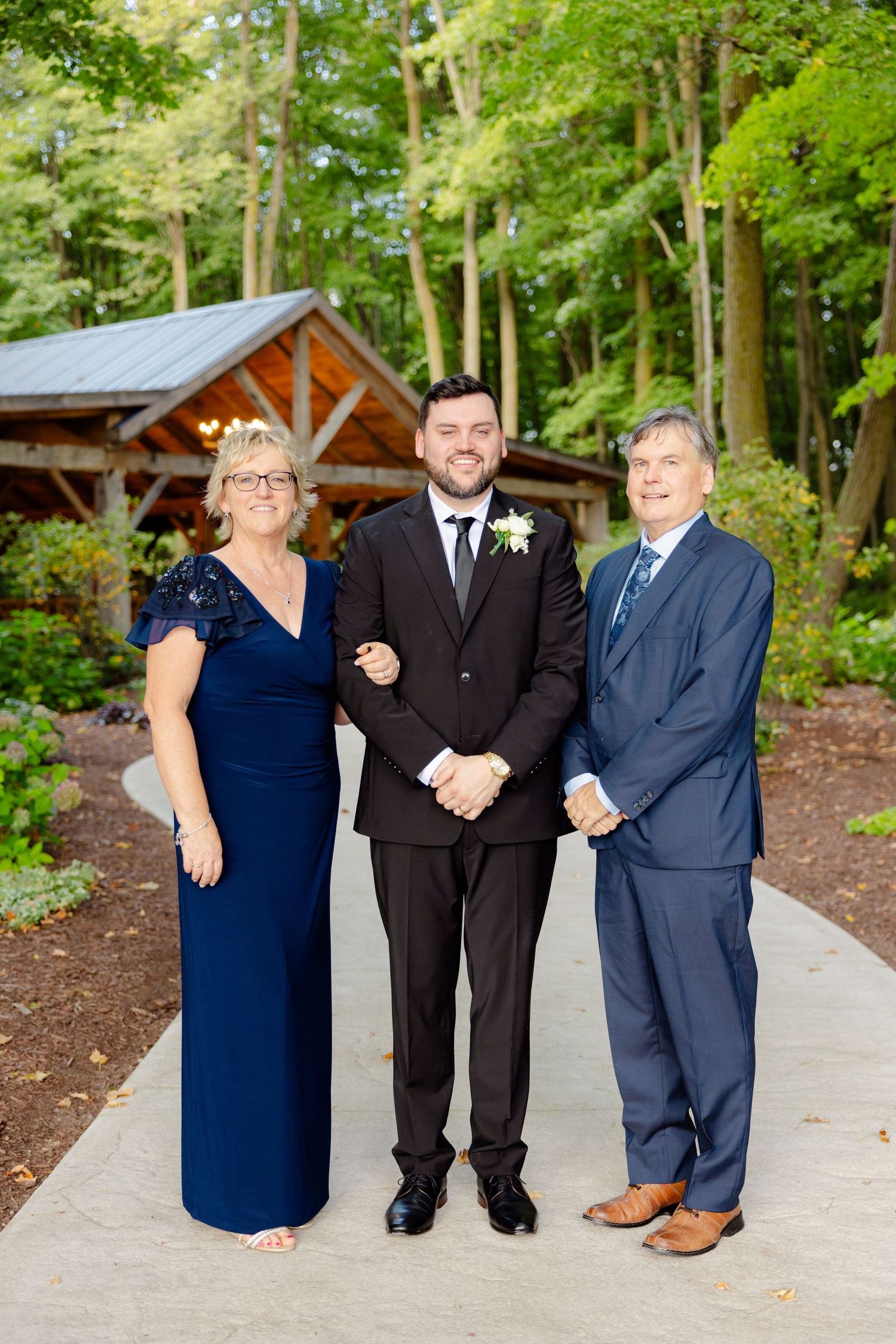 Family wedding photo showcasing three people in formal attire standing on a pathway near rustic pavilion.