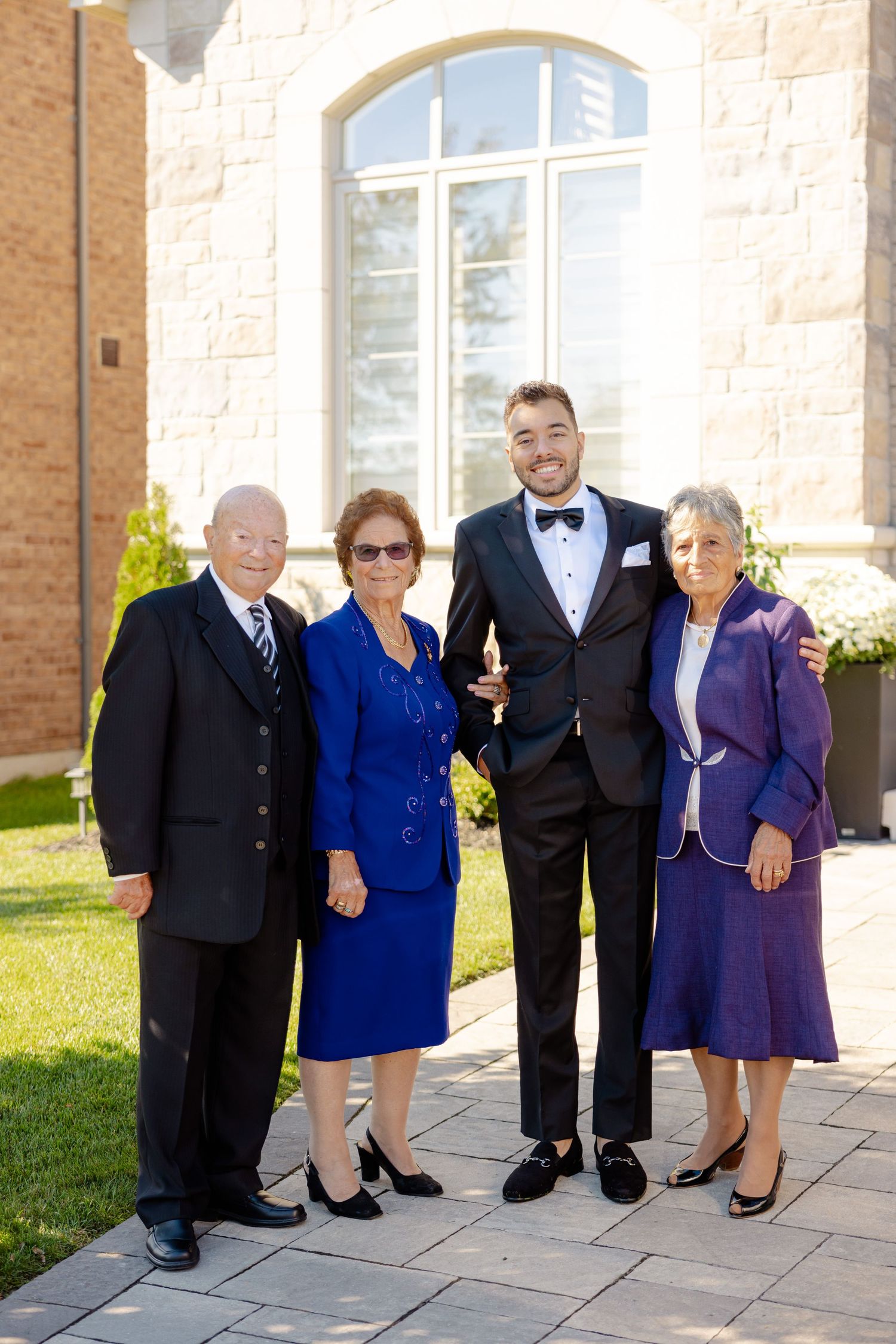 Formal wedding portrait of four people outside stone church on sunny day.