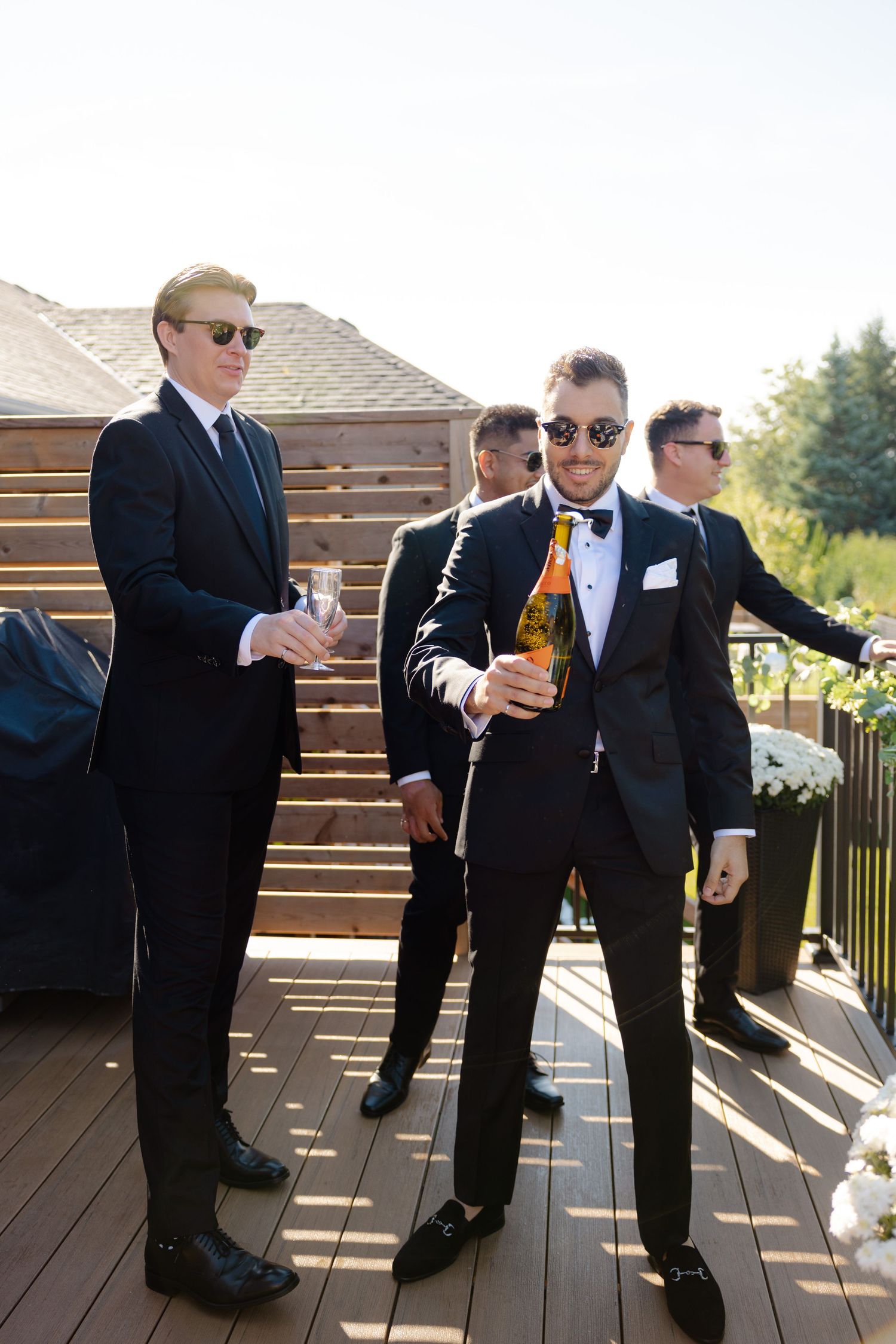 Wedding party members in sunglasses and tuxedos enjoying drinks on sunny deck.