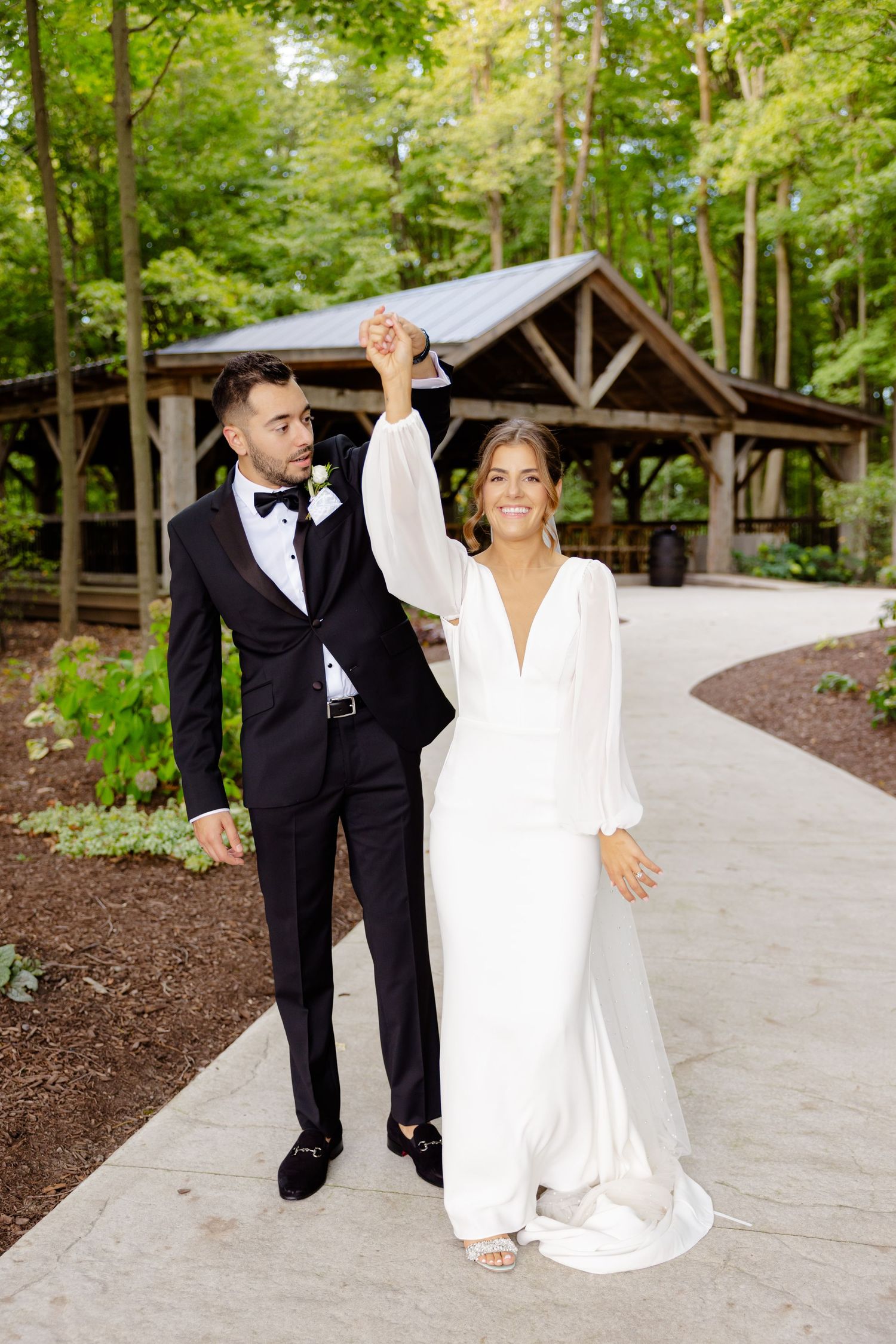 Newlyweds dance and twirl on a winding path with a rustic pavilion backdrop.
