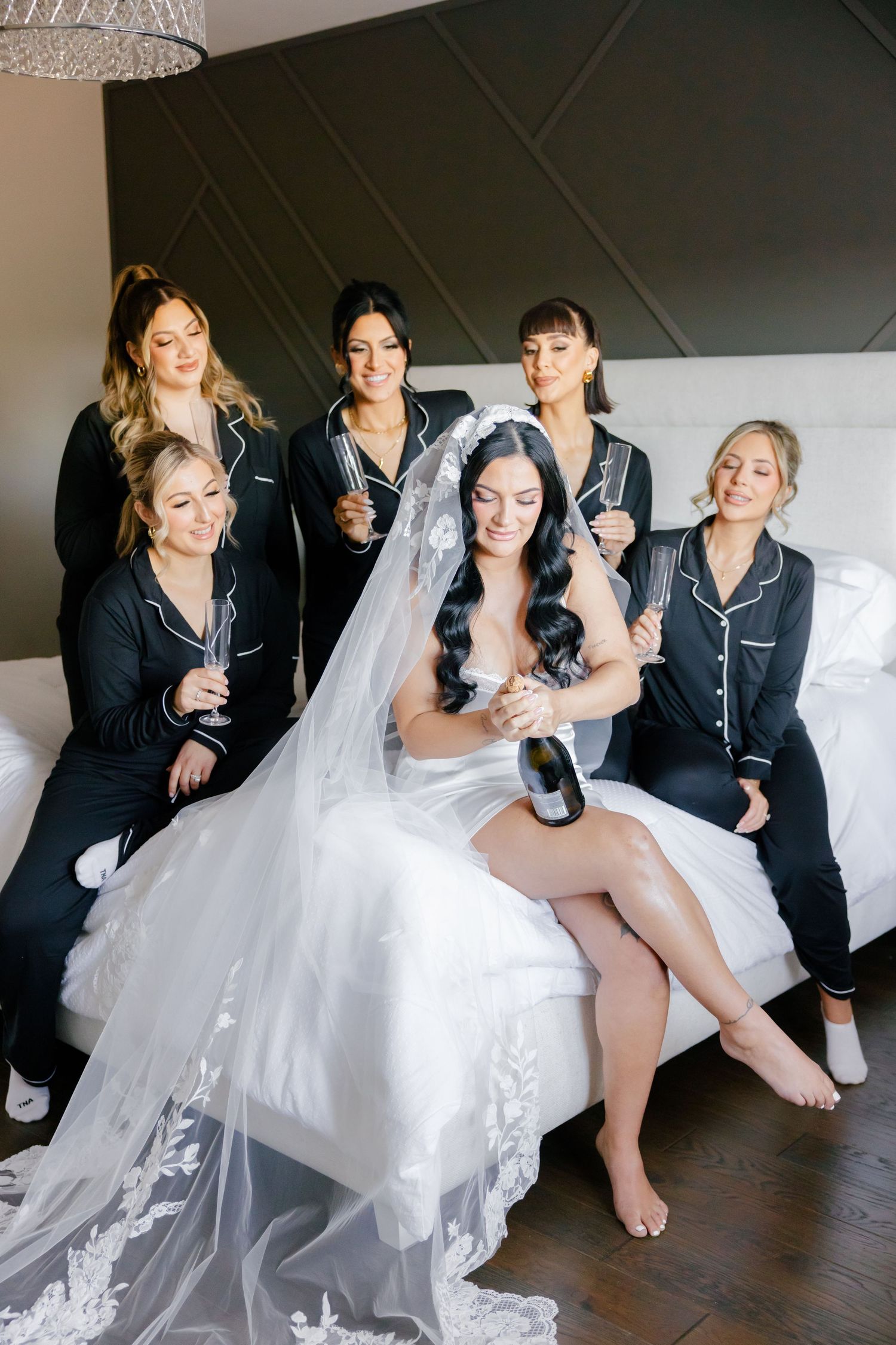 Bridesmaids in matching black pajamas celebrate with bride in white gown on bed.