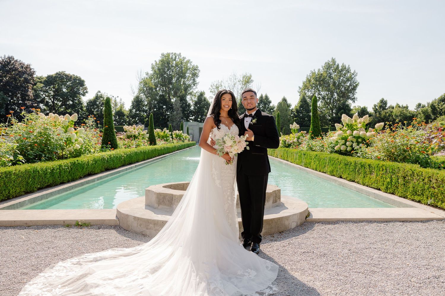 A couple in wedding attire poses by a garden fountain surrounded by manicured hedges and flowers.