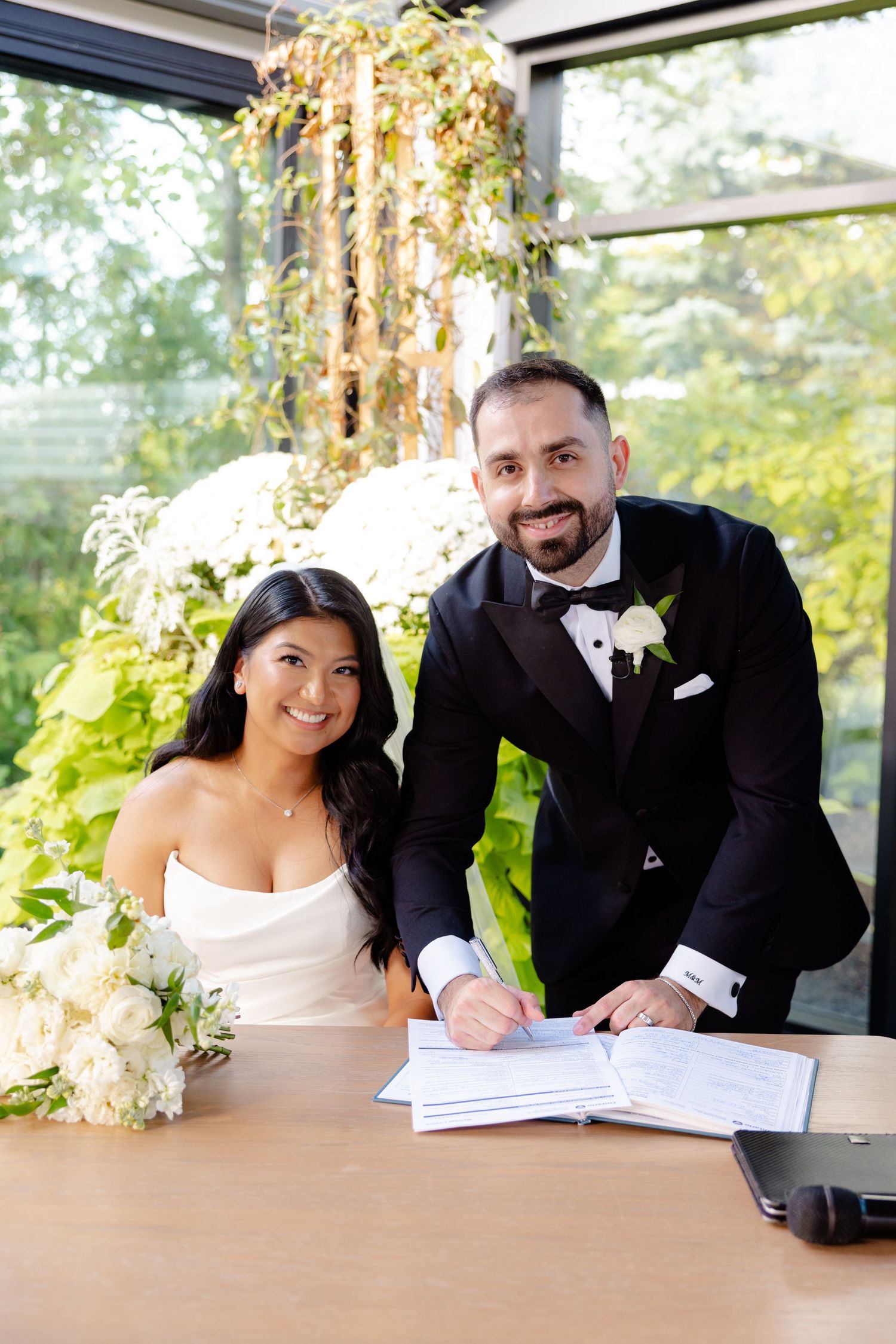 A couple signs wedding documents together at a wooden table with decorative greenery behind them.