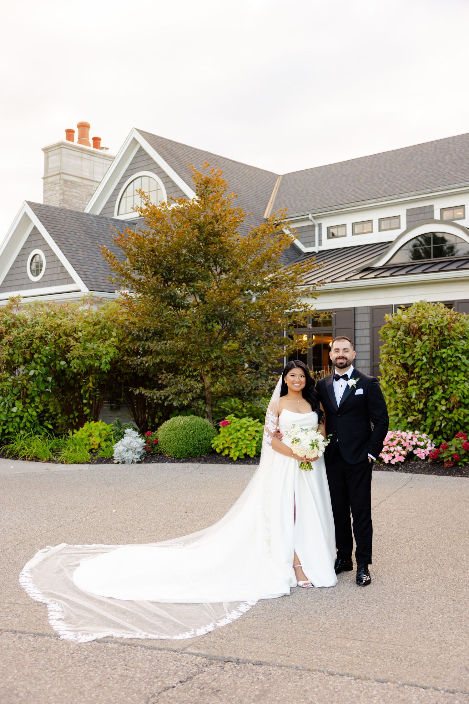 A wedding couple poses in front of a beautiful grey building with fall foliage and landscaping.