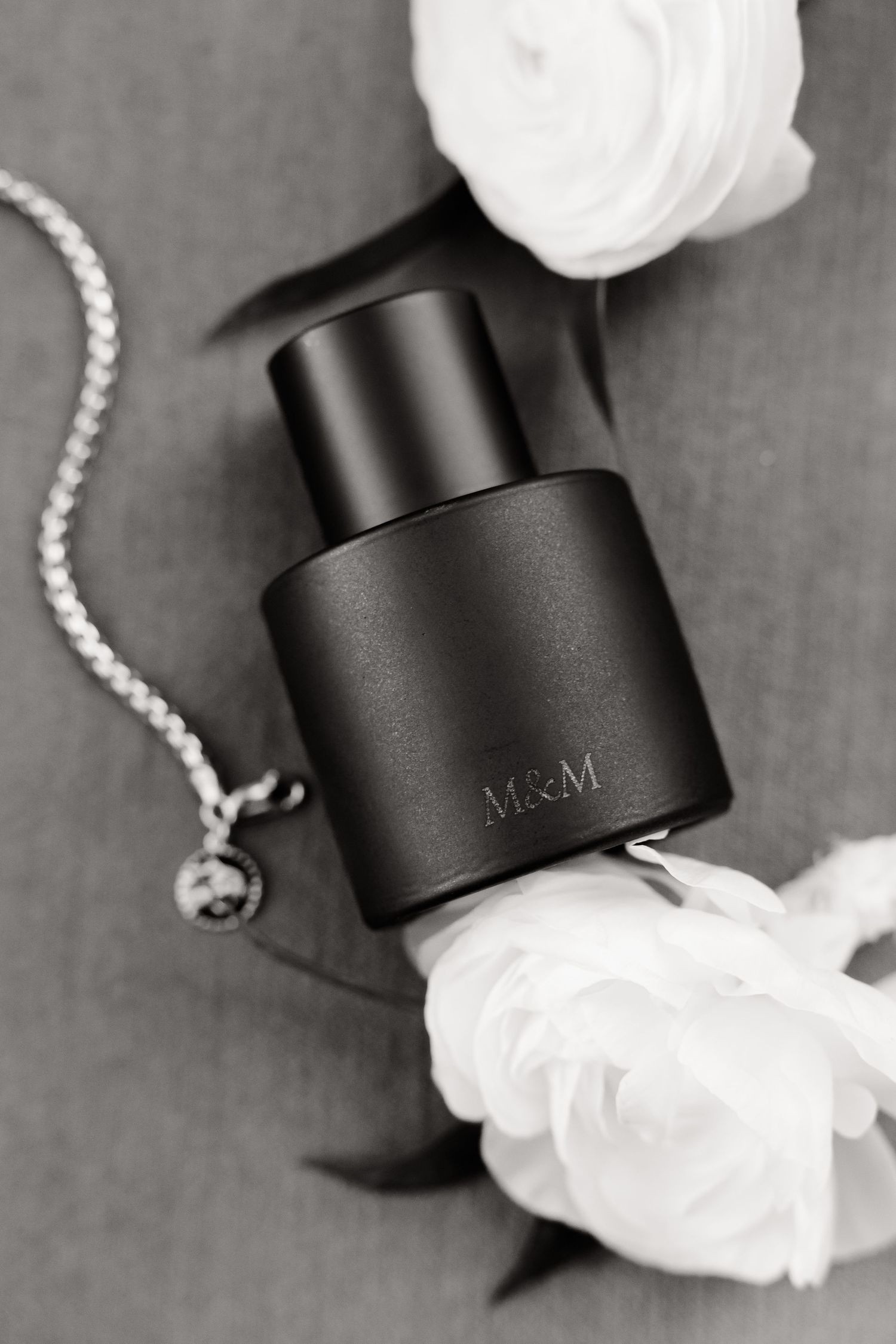 Black M&M perfume bottle styled with white roses and silver chain in elegant monochrome composition.