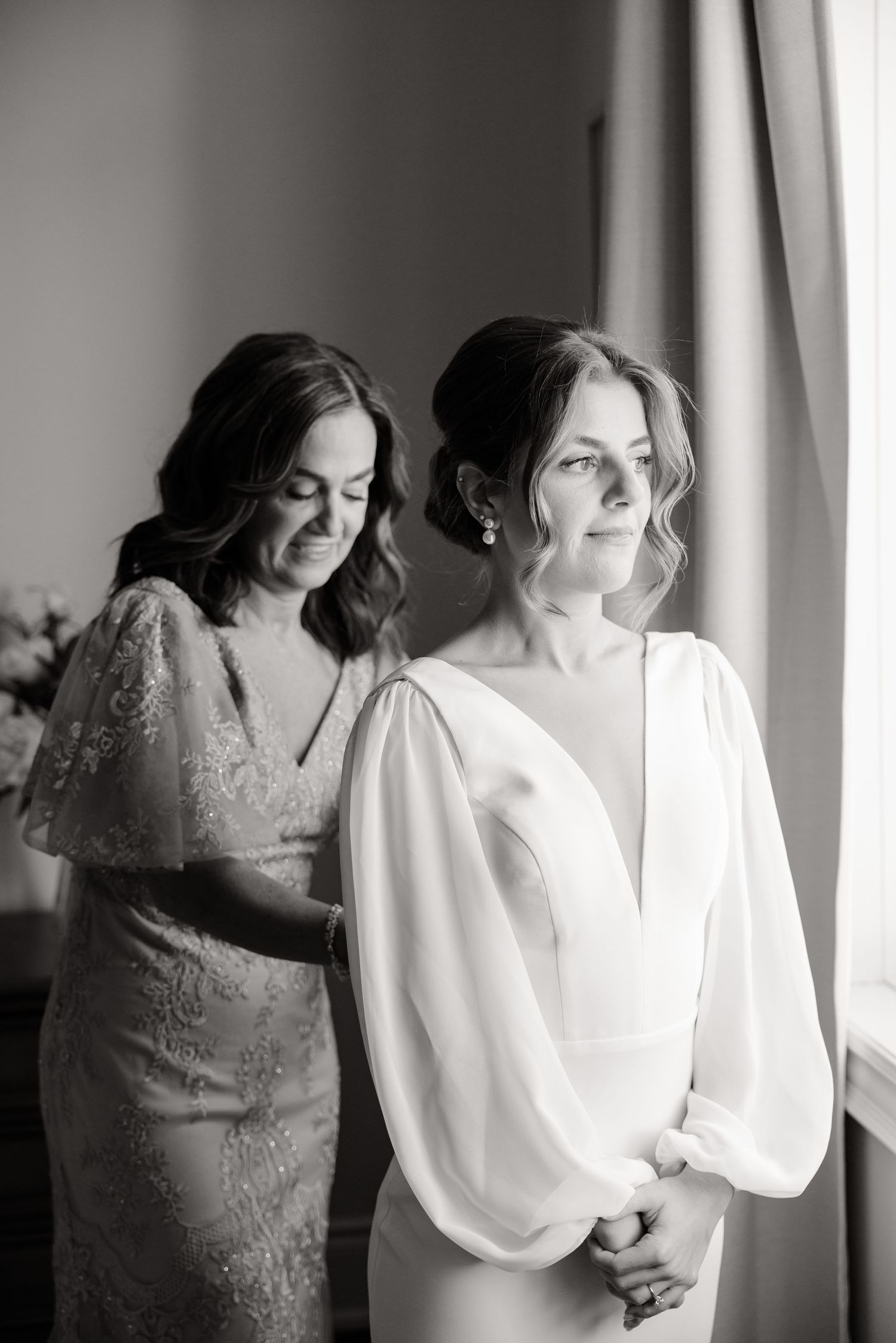 Two people in formal dresses stand by a window, one in white and one in beige lace.