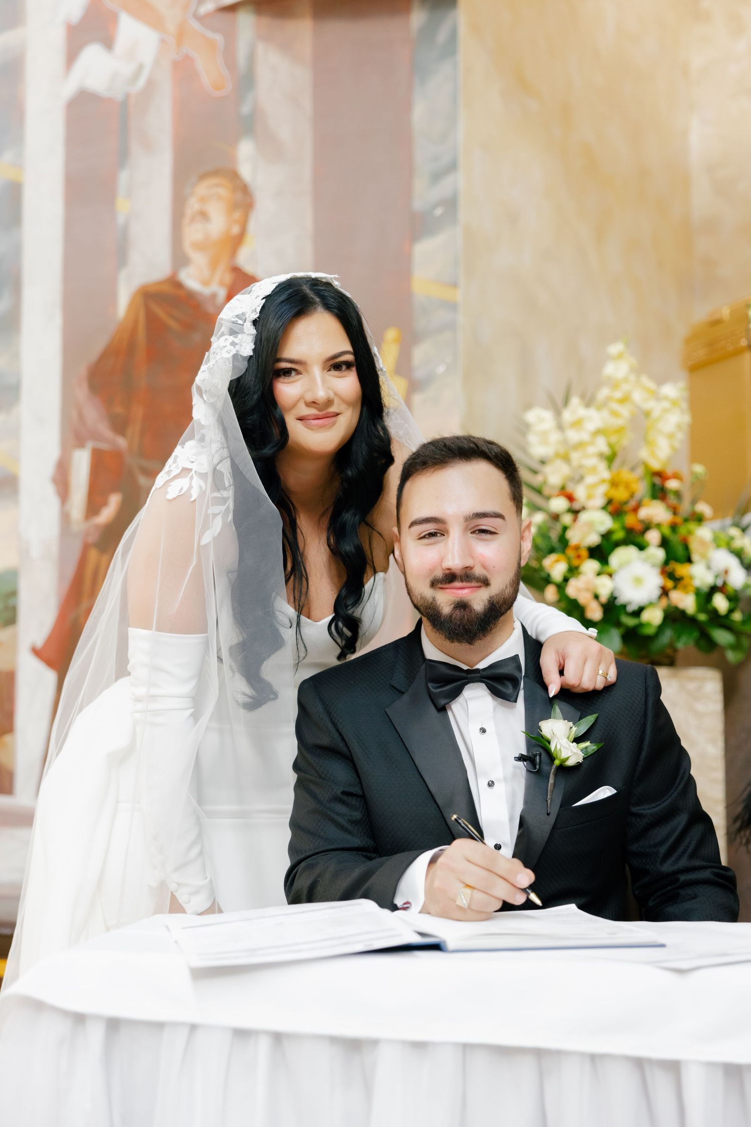 Newlyweds pose while signing marriage documents at a decorated table.