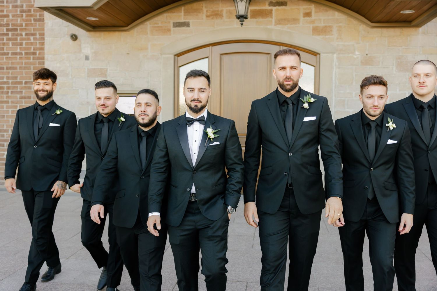 A group of groomsmen in black suits pose together in front of a beige brick building with an arched doorway.