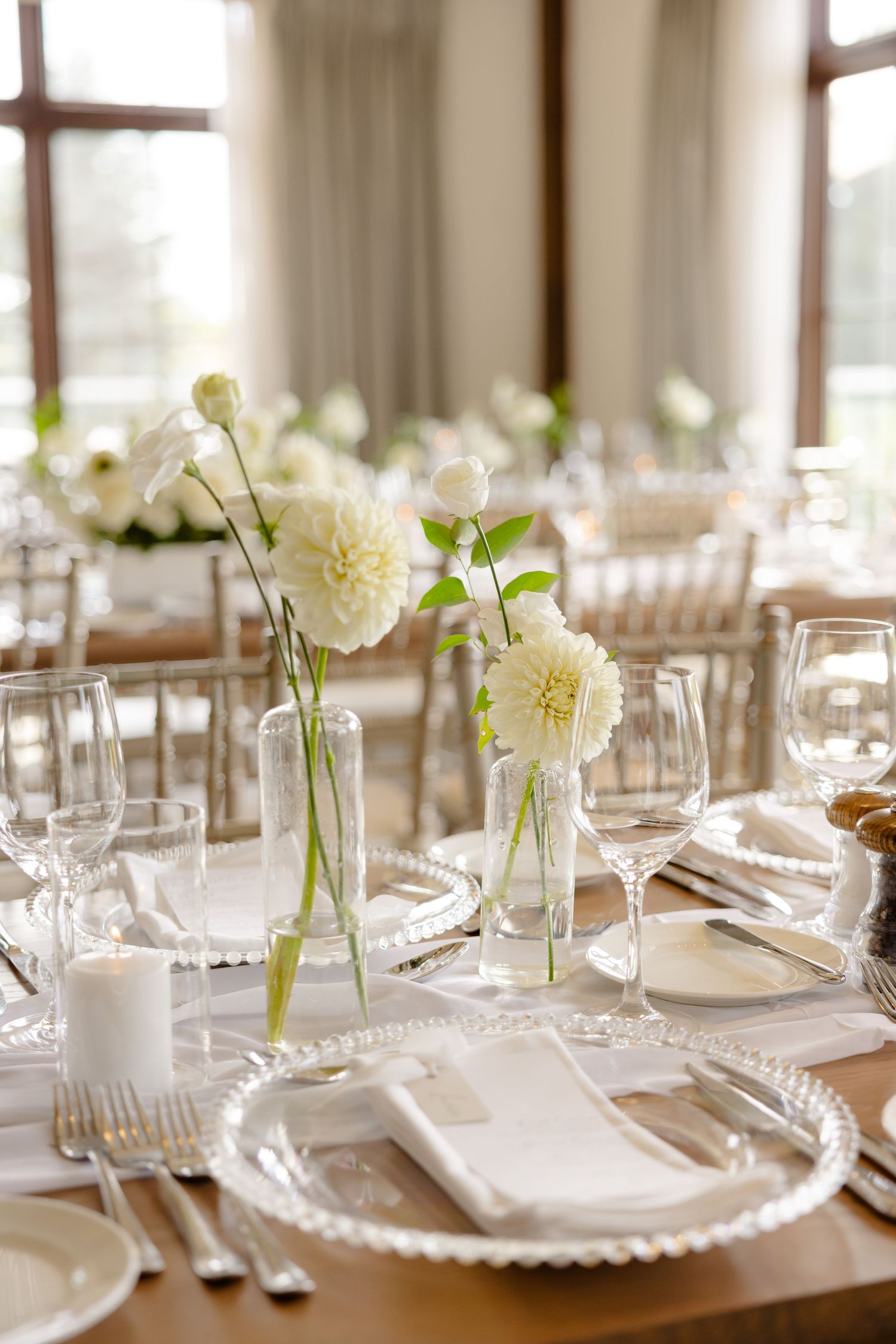 Bright wedding reception table with white dahlia centerpieces in glass vases and elegant place settings.
