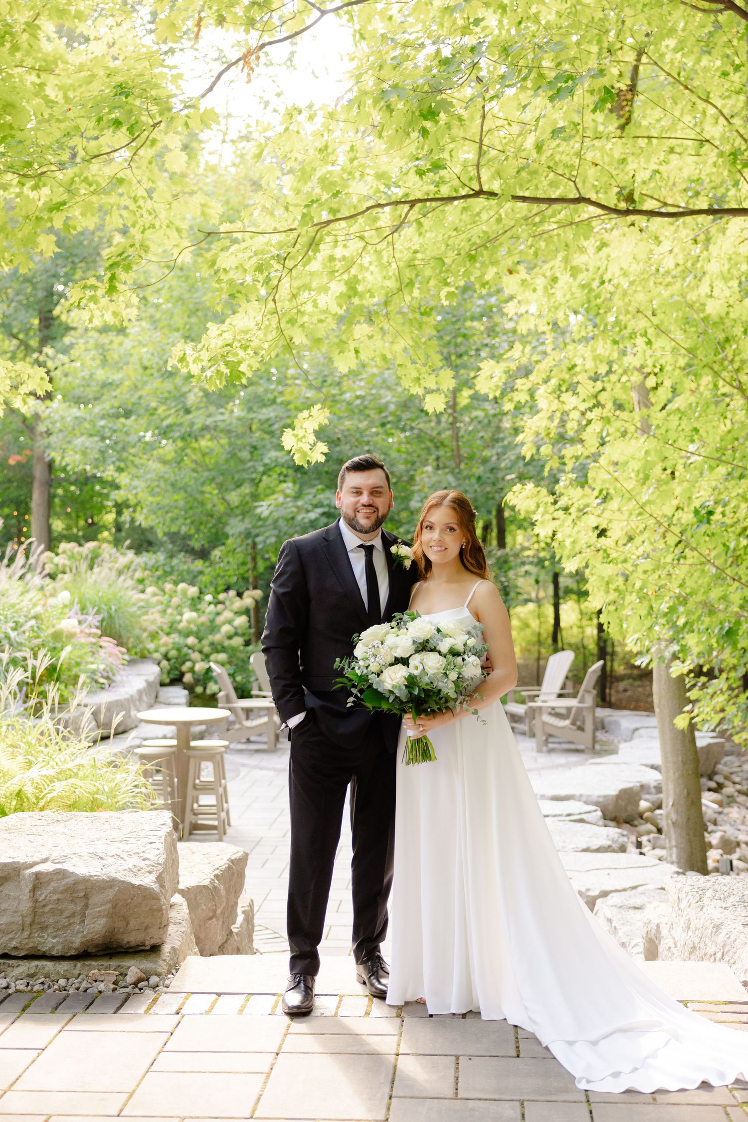 Wedding couple stands on stone path holding white floral bouquet under green canopy.