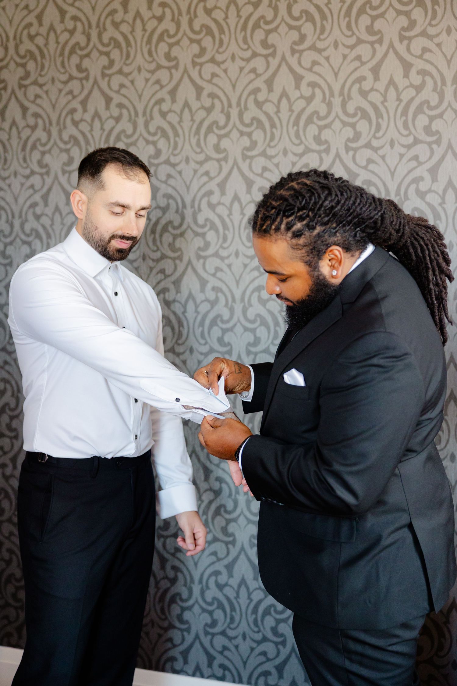 Two people in formal attire helping with cufflinks against patterned wallpaper background.