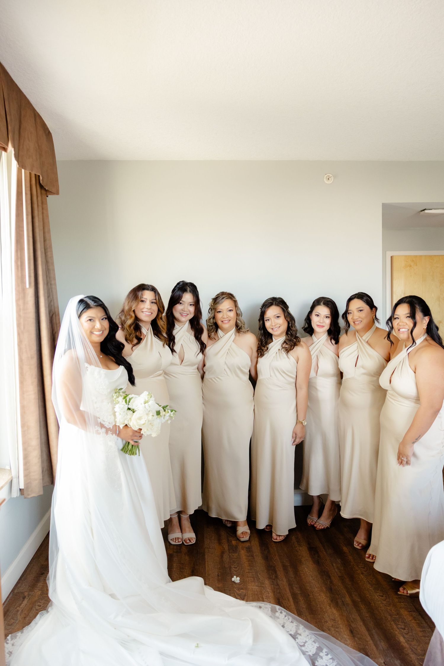 Bridal party in coordinated cream dresses stands by a window for a formal wedding photo.