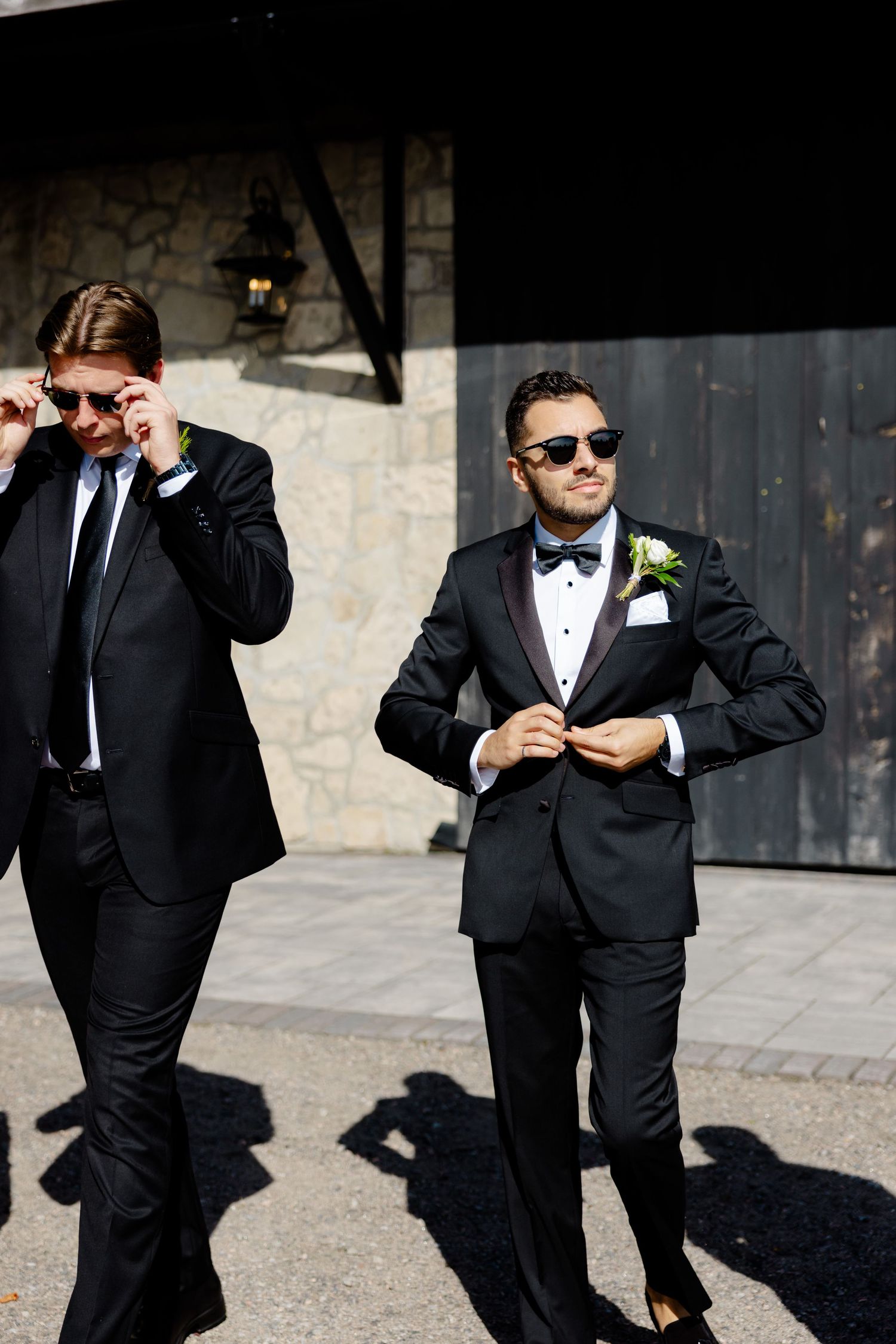 Two groomsmen in classic black tuxedos and sunglasses pose against an industrial stone wall.