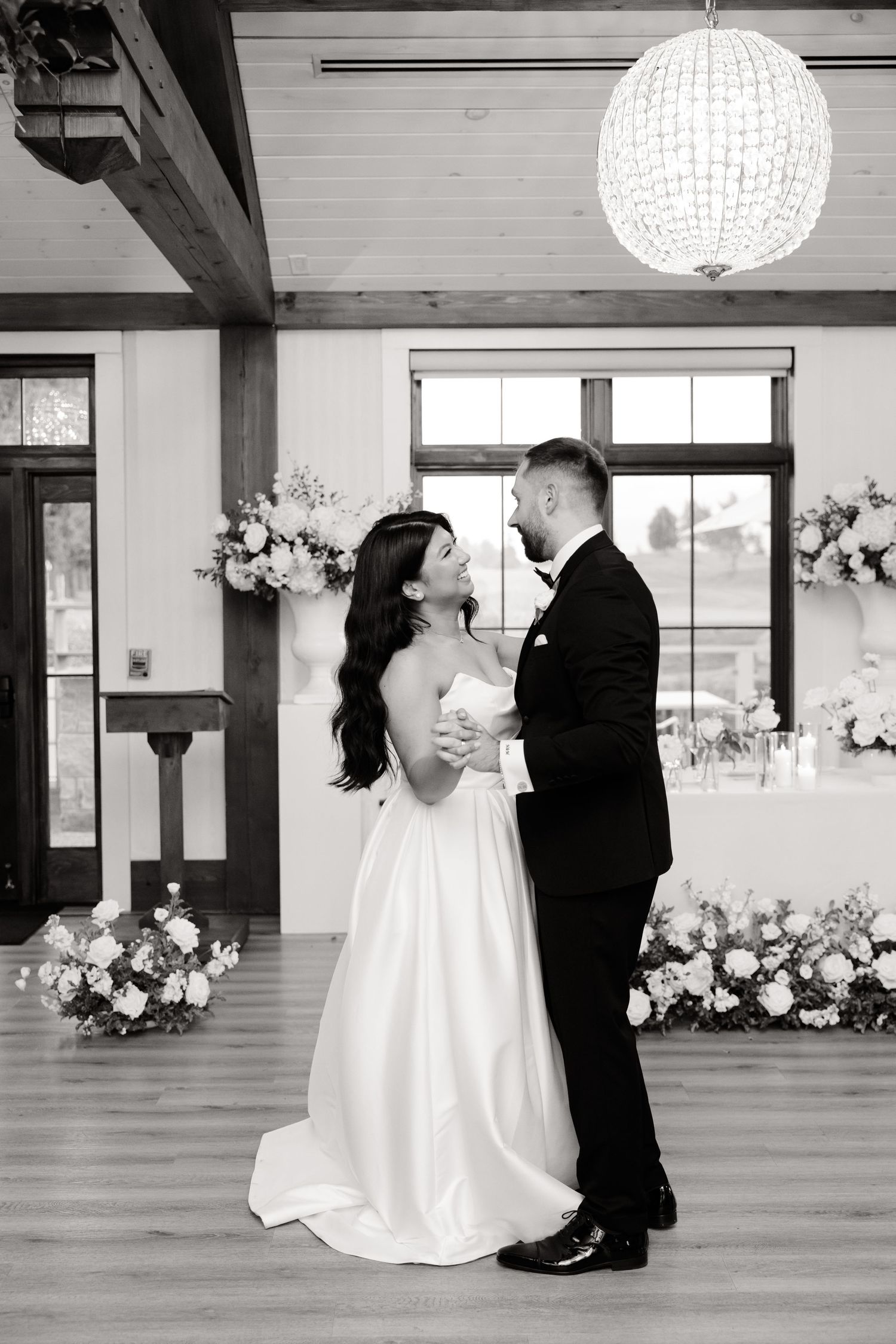 Black and white photo of a wedding dance in a venue with rustic wooden beams and floral decor.