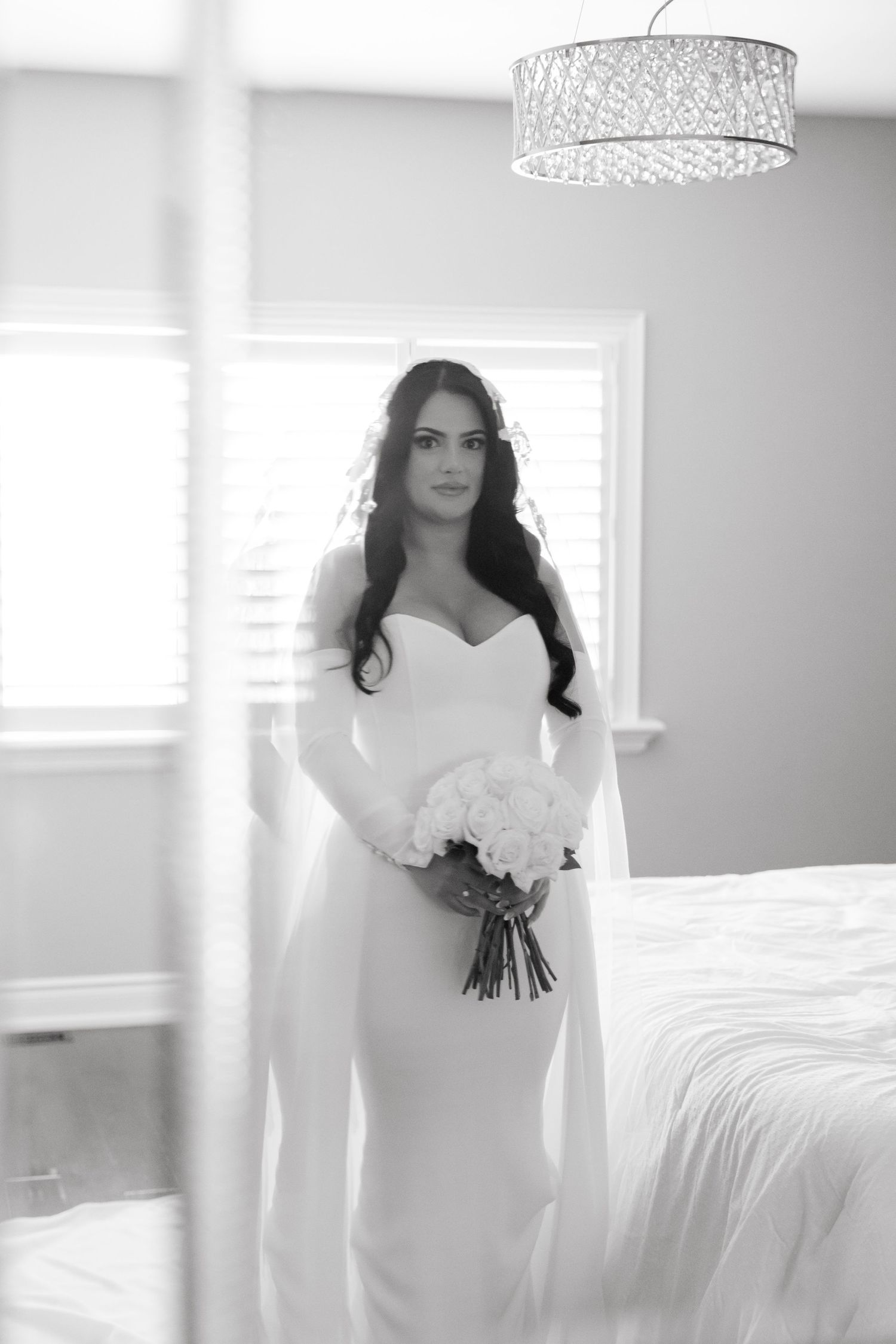 Black and white bridal portrait with crystal chandelier in bright bedroom setting.