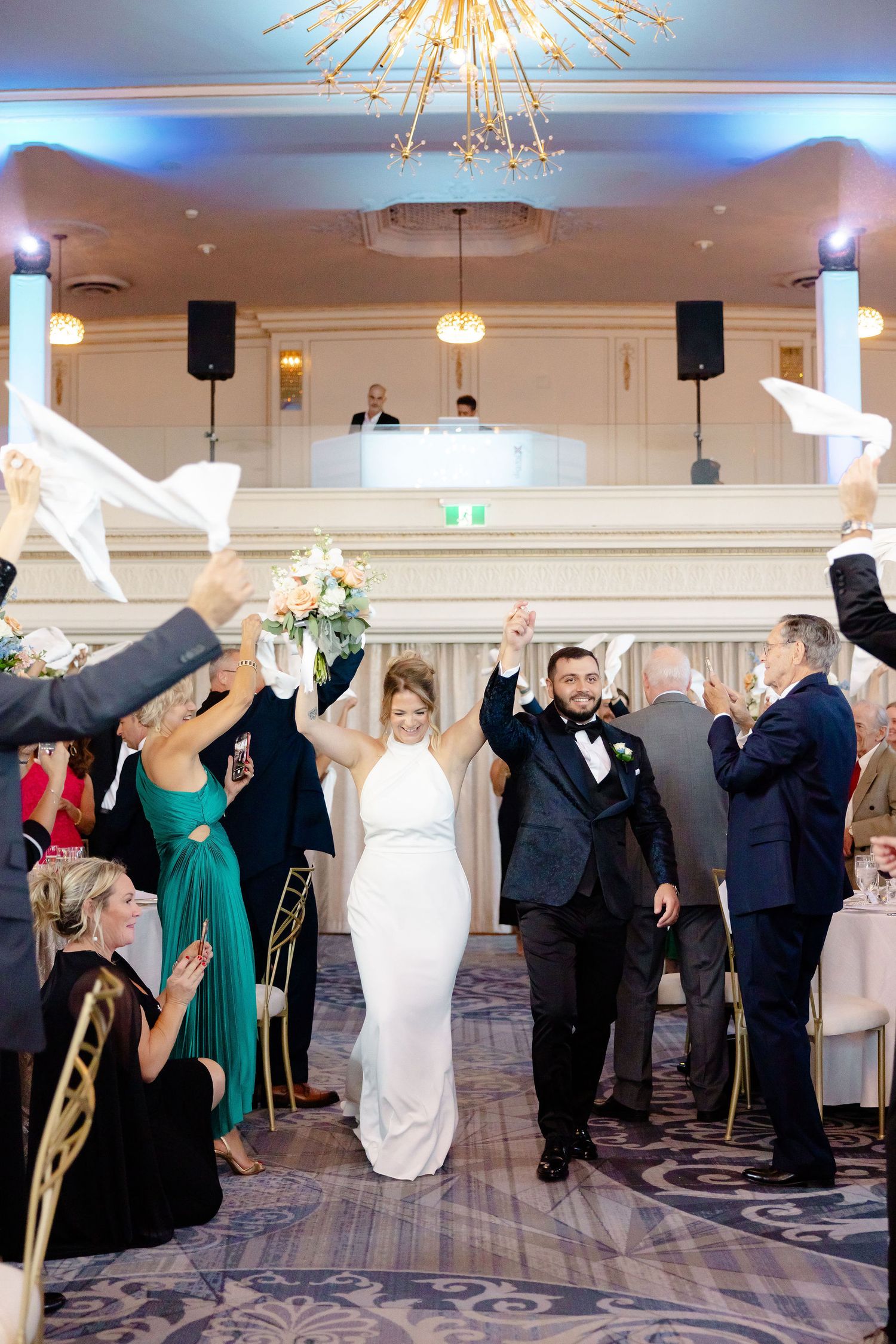 Wedding party members raise their arms in celebration during a reception under a grand chandelier.