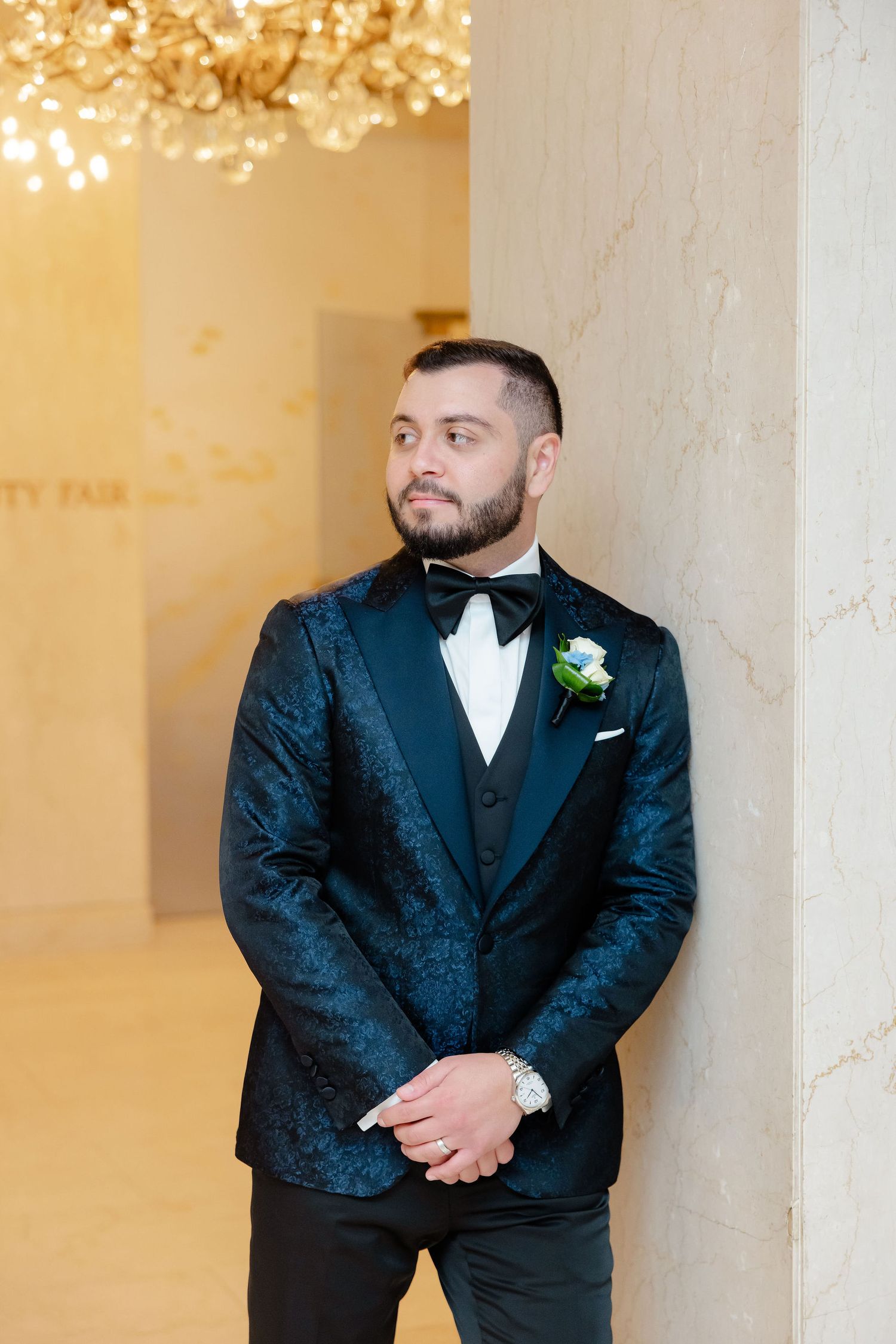 Groom in textured black tuxedo poses against cream-colored wall under crystal chandelier lighting.
