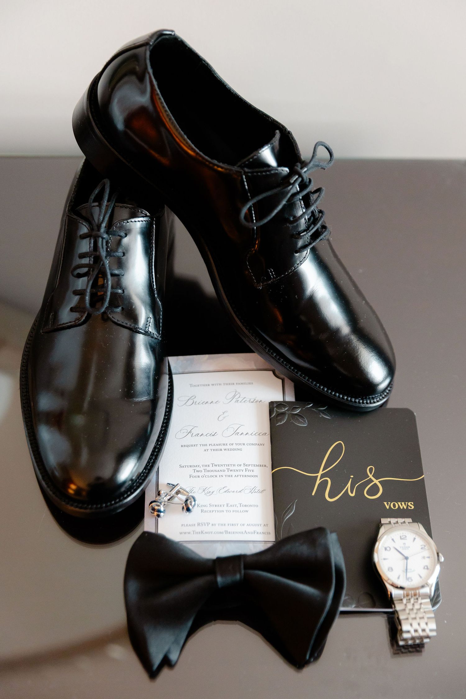 Black leather dress shoes displayed with a watch and invitation card on a gray surface.