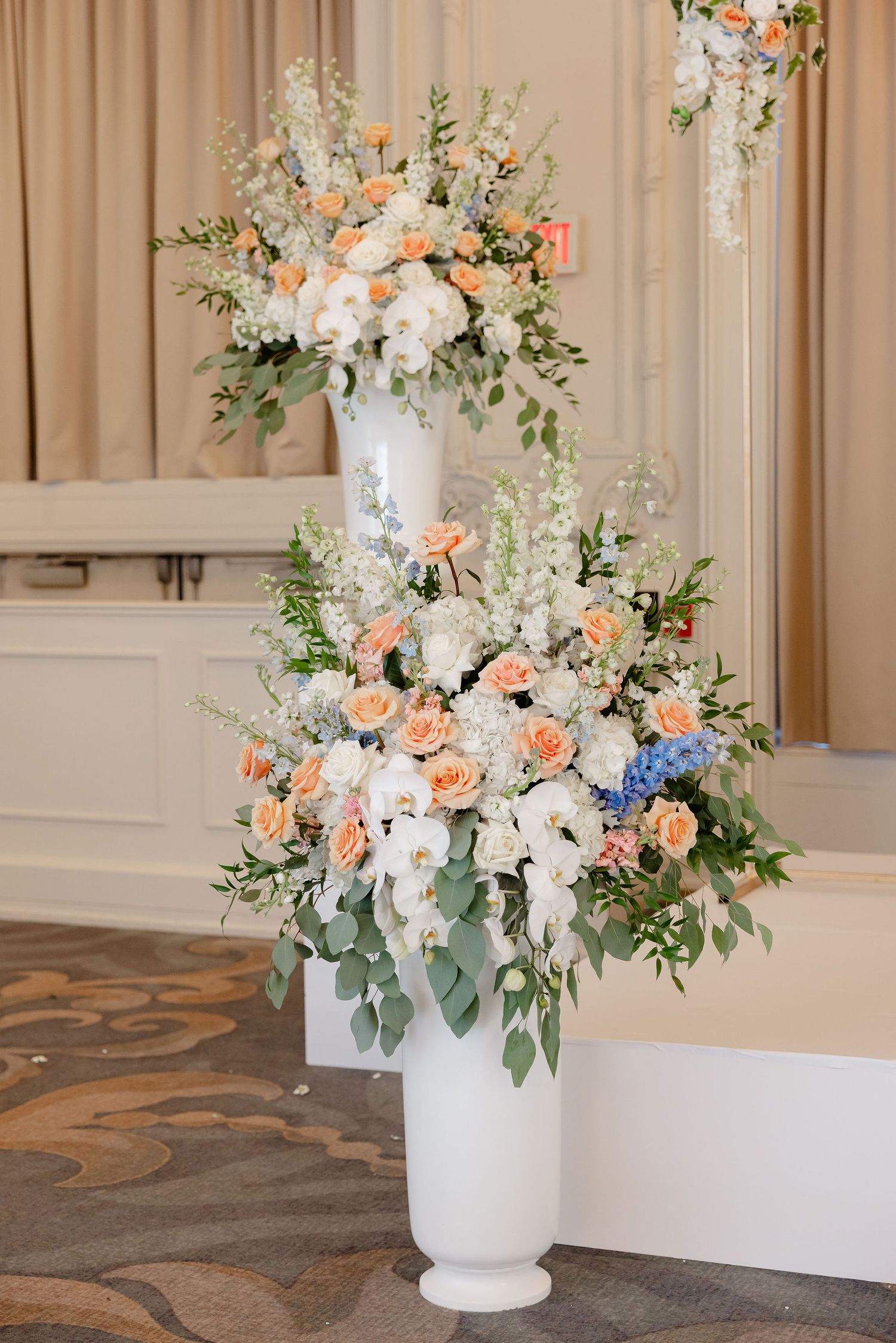 Tall white pedestal vase with lush peach, white and blue floral arrangement featuring roses, hydrangeas and cascading greenery.