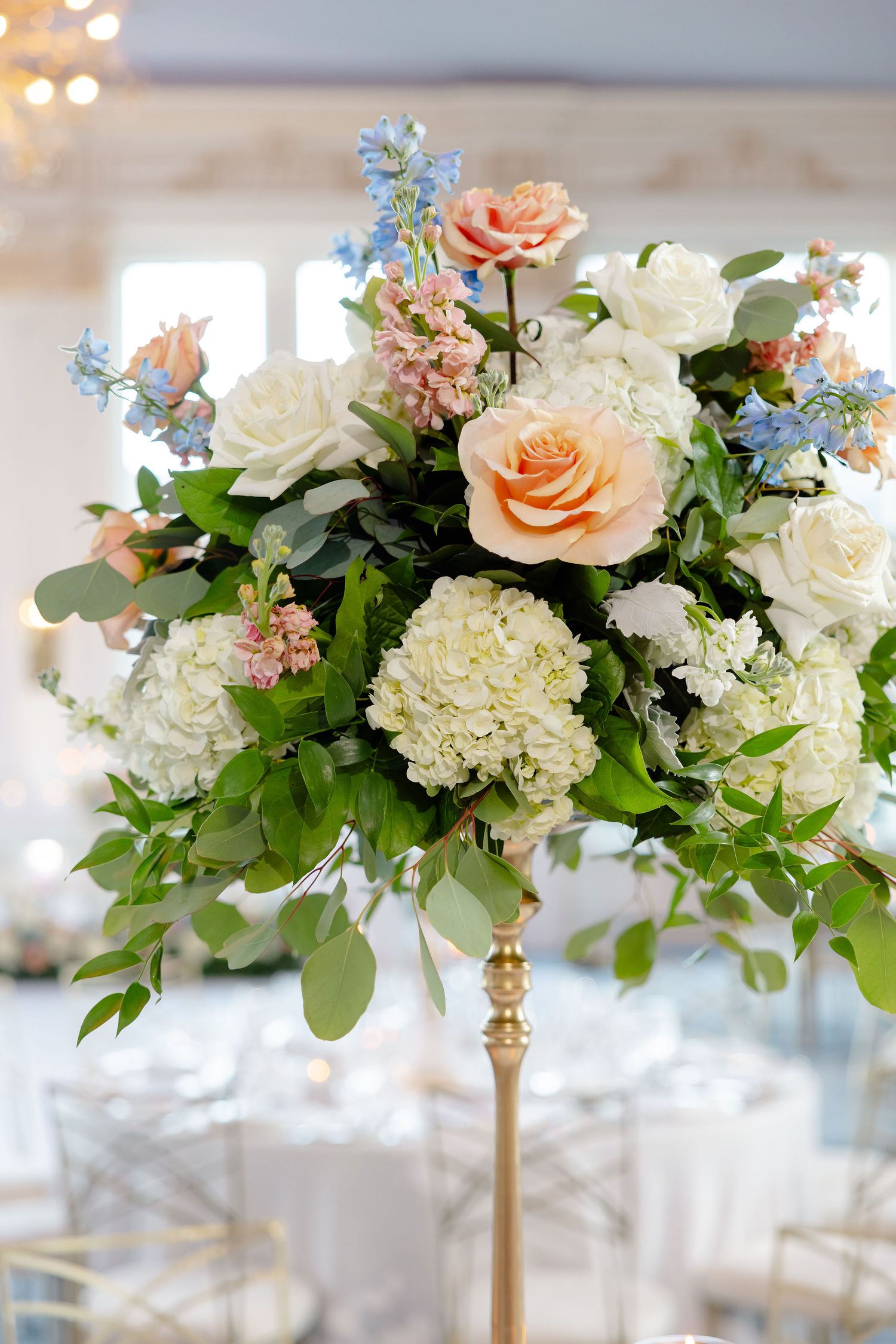 Elegant floral centerpiece with peach roses, white hydrangeas, and blue delphinium on a gold pedestal stand.