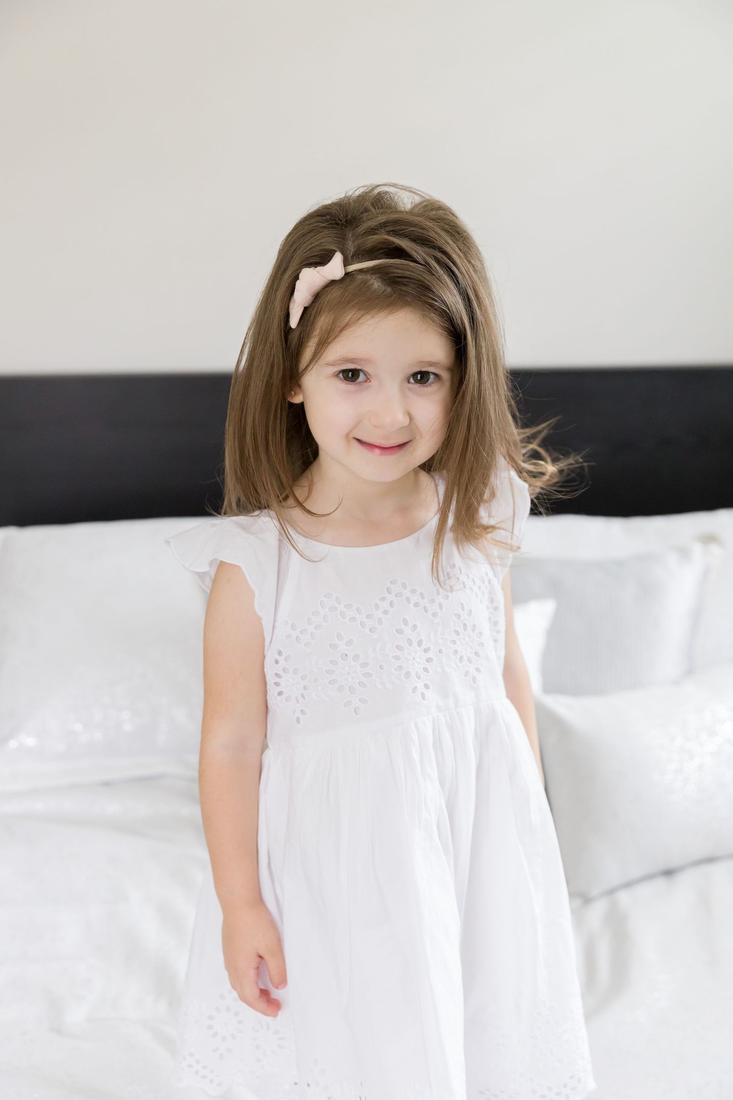 Young child wearing white eyelet dress stands on bed with white linens and dark headboard.