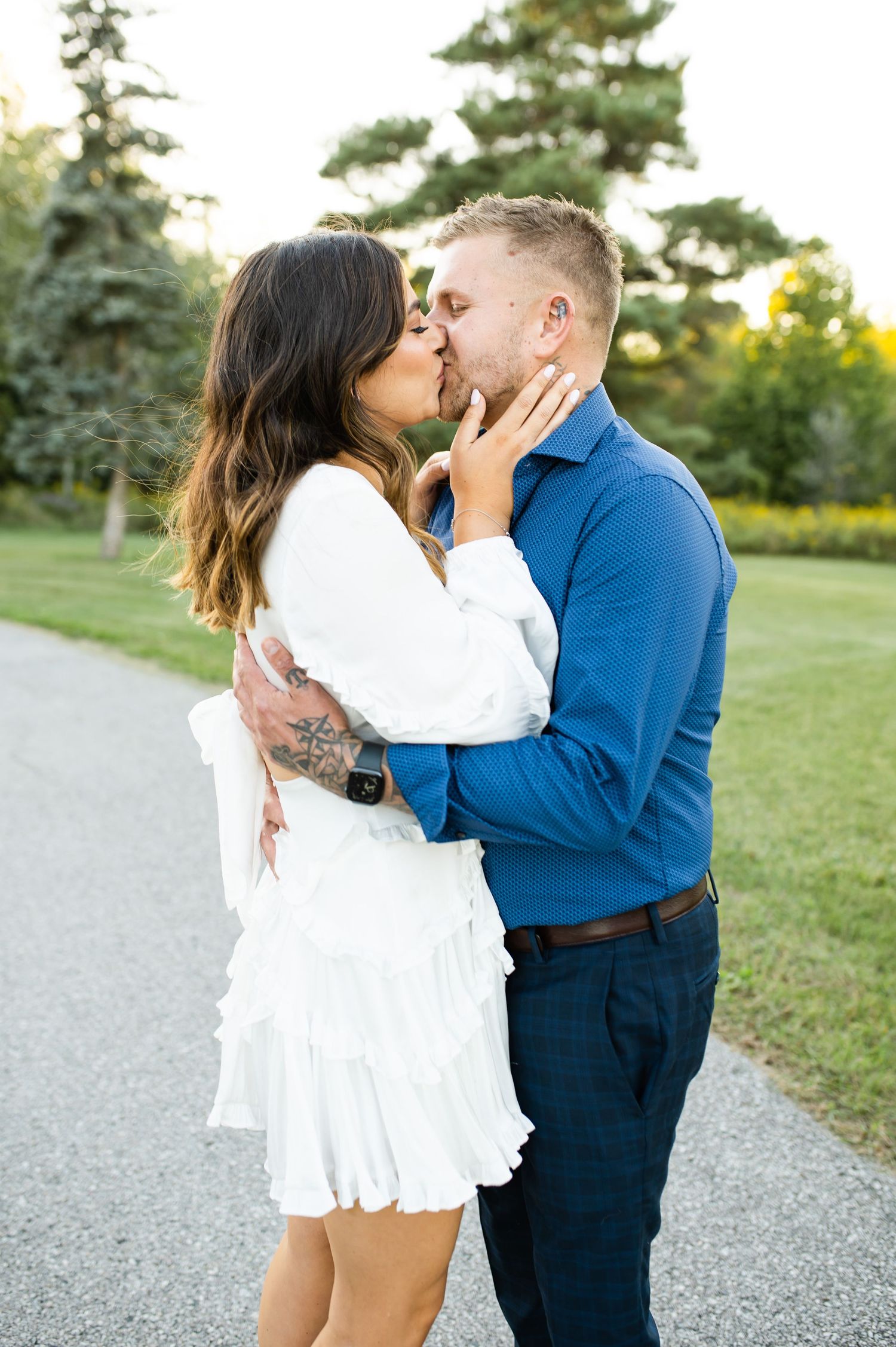 Couple sharing romantic kiss on garden path with evergreen trees in background.
