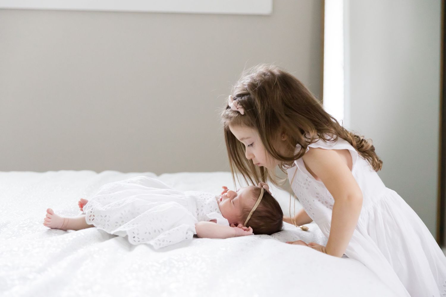 Young child leaning over to look at sleeping baby on white bed.