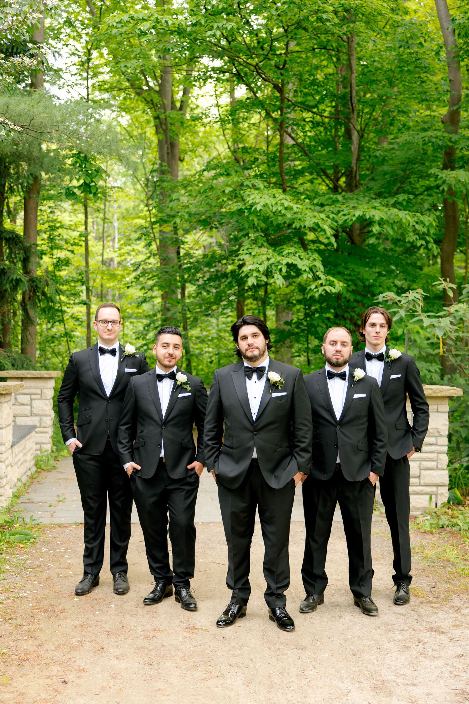 Groomsmen in black tuxedos posing on stone path in lush forest setting.