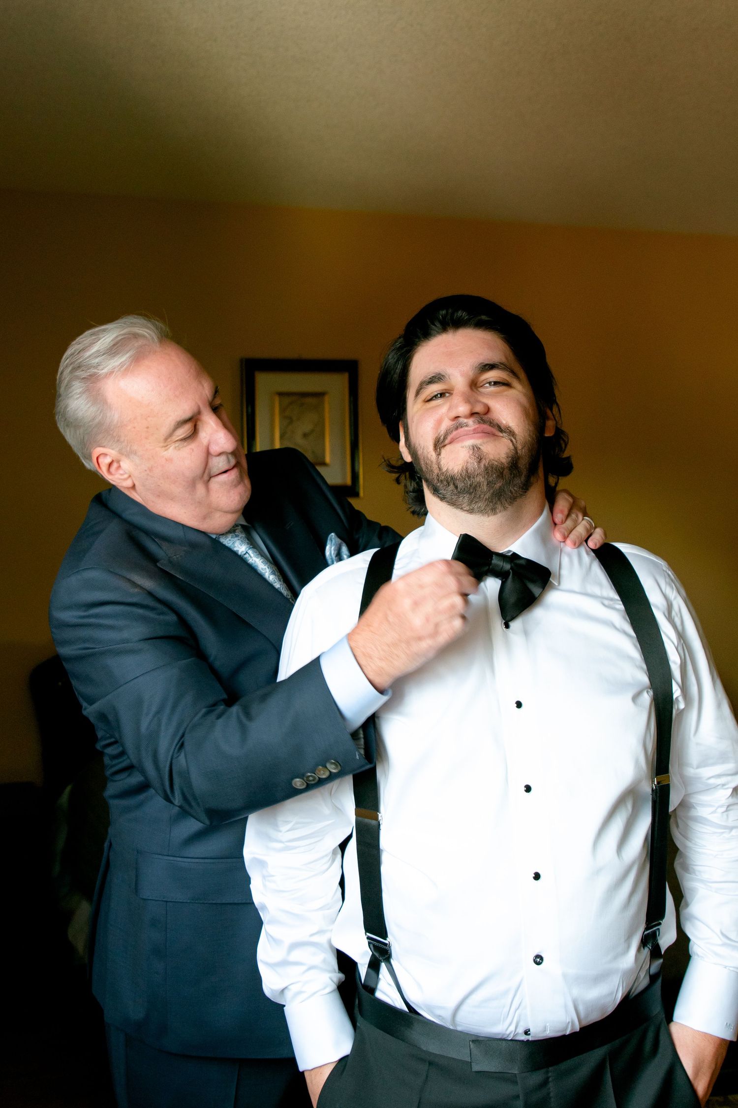Two men in formal attire sharing a moment while adjusting a bowtie before an event.