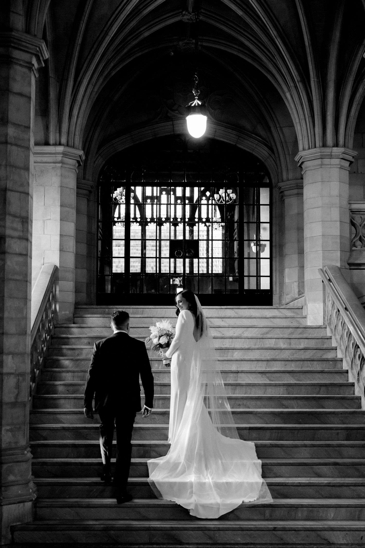 A romantic black and white photo of newlyweds ascending a grand stone staircase.