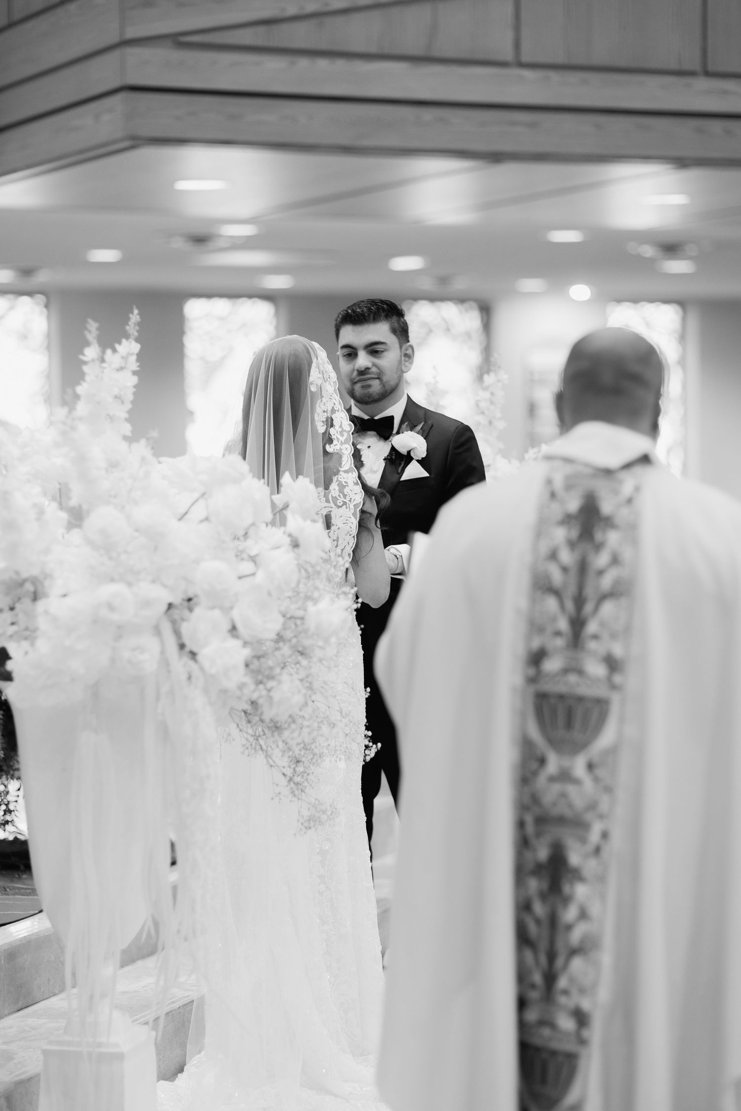 Black and white photo of a wedding ceremony with priest officiating in front of decorative floral arrangements.