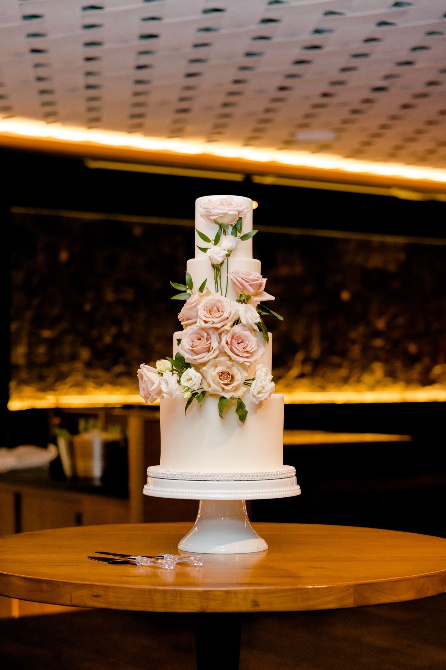 Elegant white wedding cake decorated with pale pink roses and greenery on wooden table.