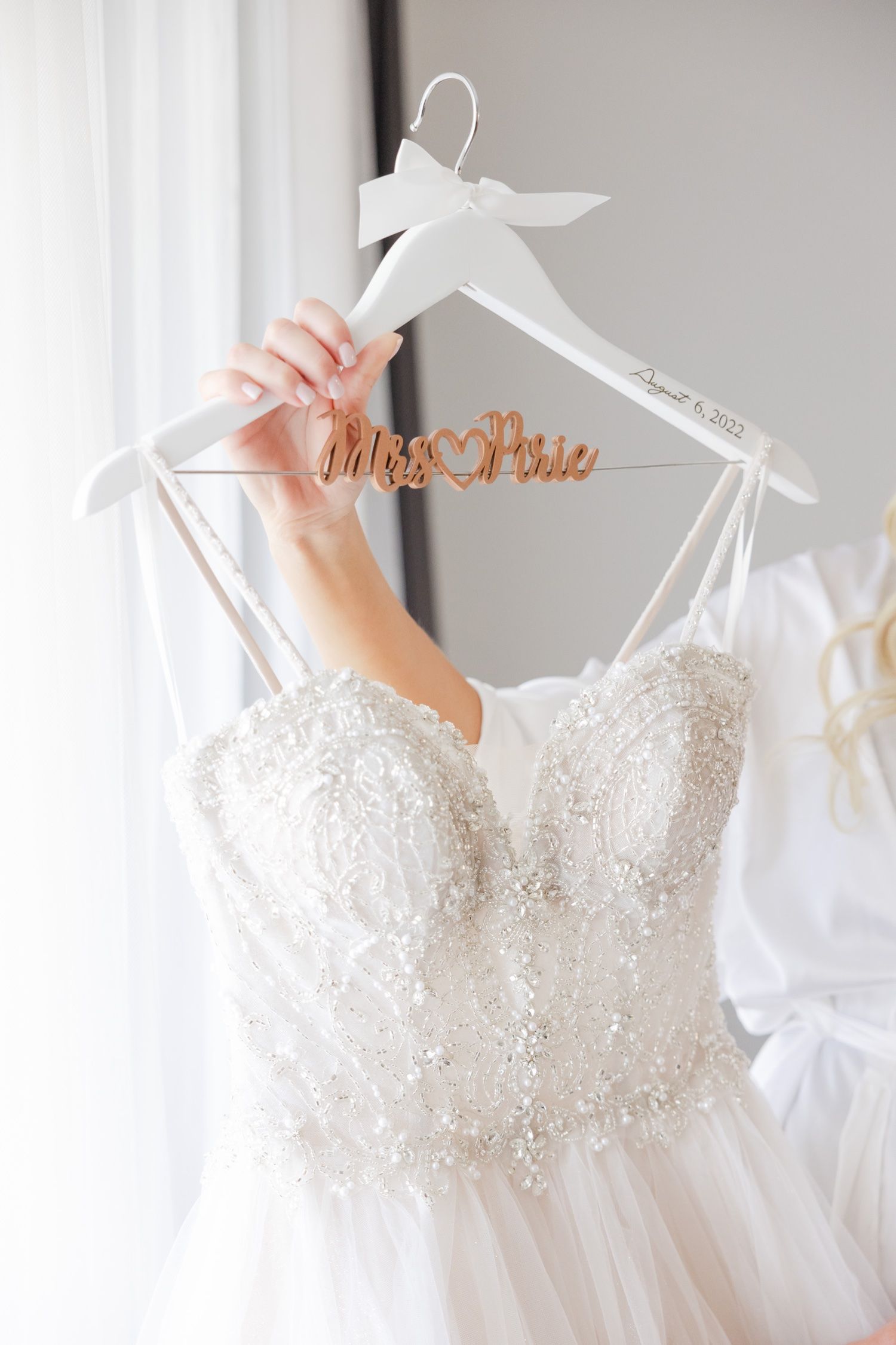 A beautiful white wedding dress with intricate lace detailing hangs on a personalized hanger against a grey wall.