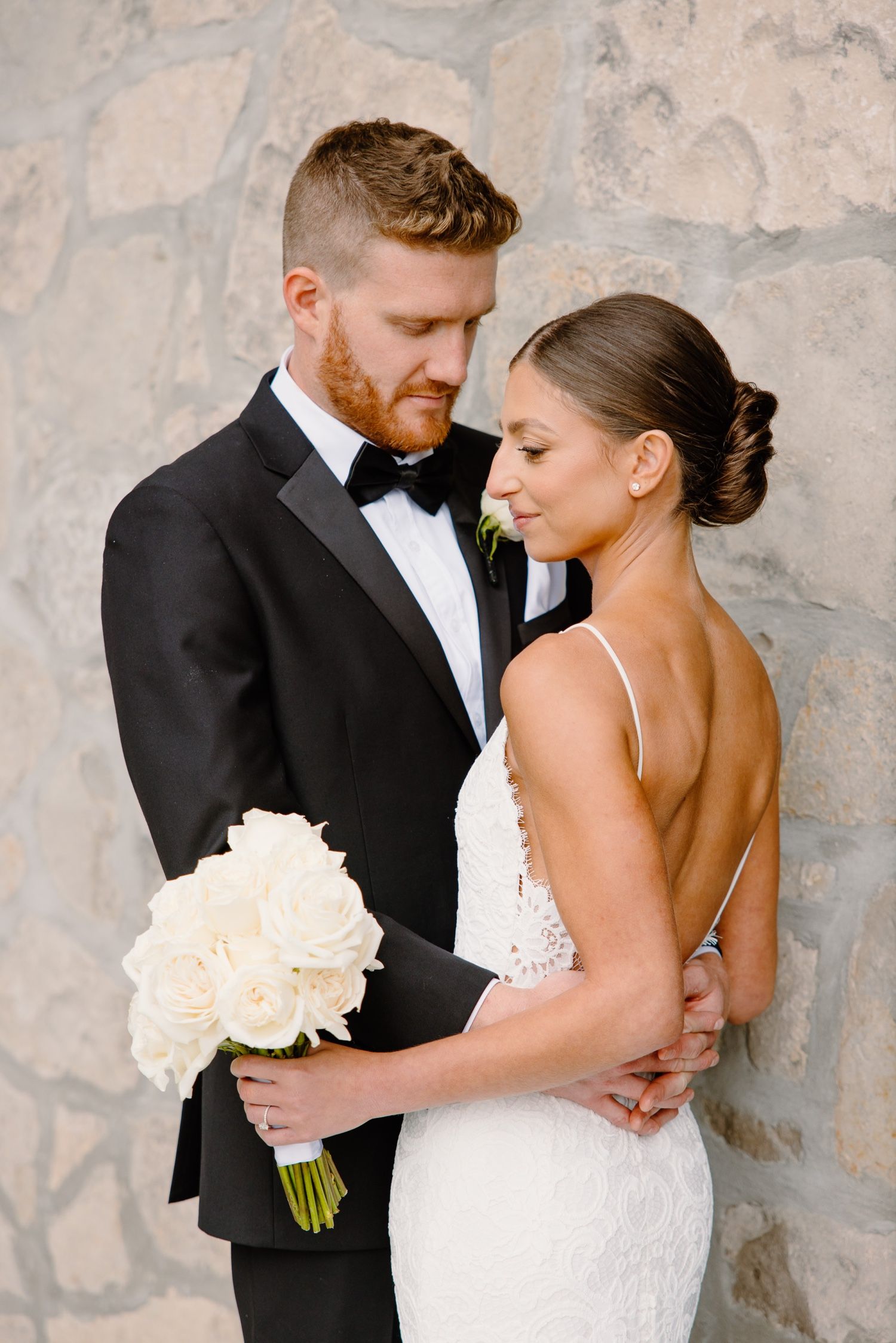 Tender moment captured against textured stone wall with white rose bouquet.