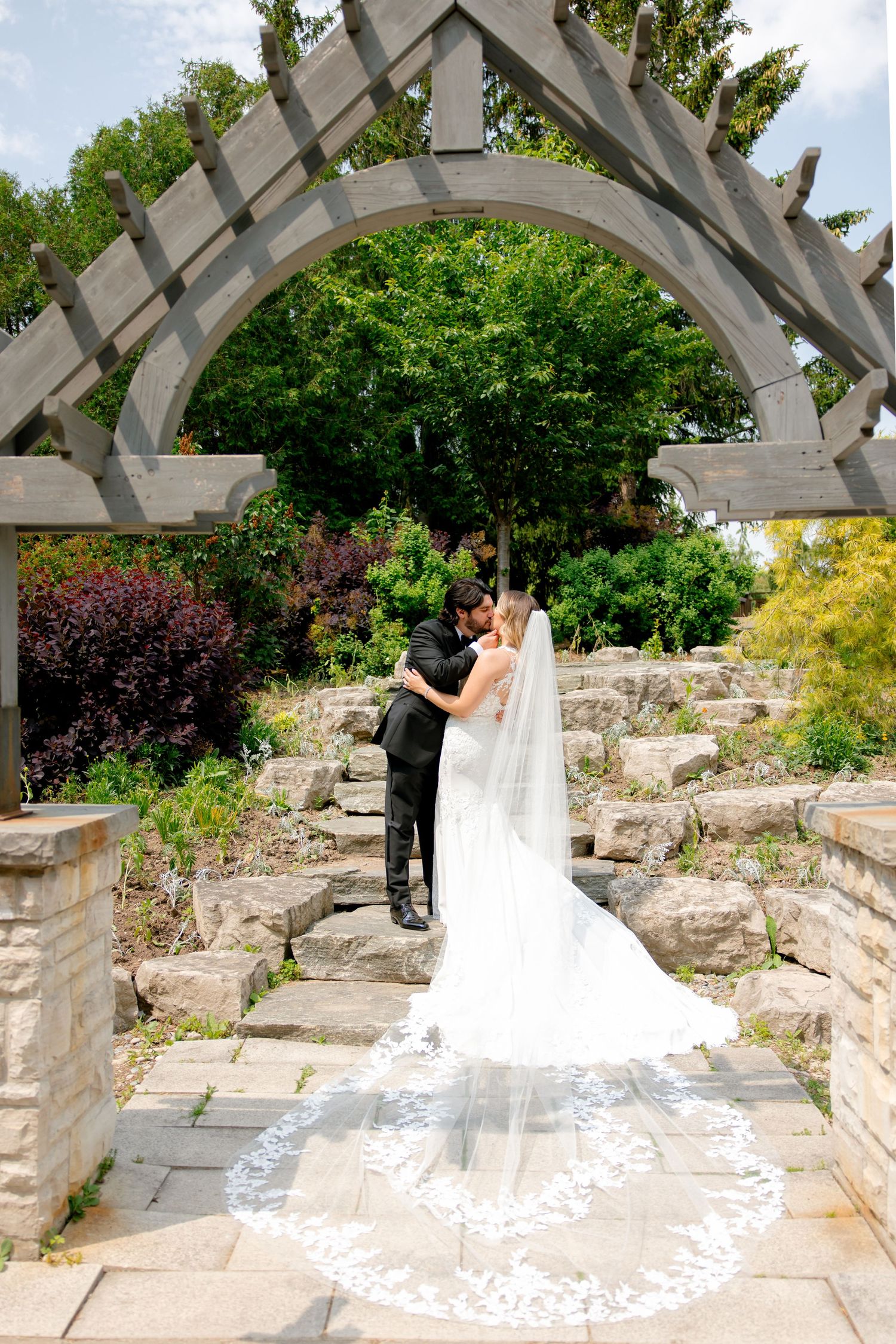 A couple shares a romantic moment under an elegant garden archway with a stunning long veil trailing down stone steps.