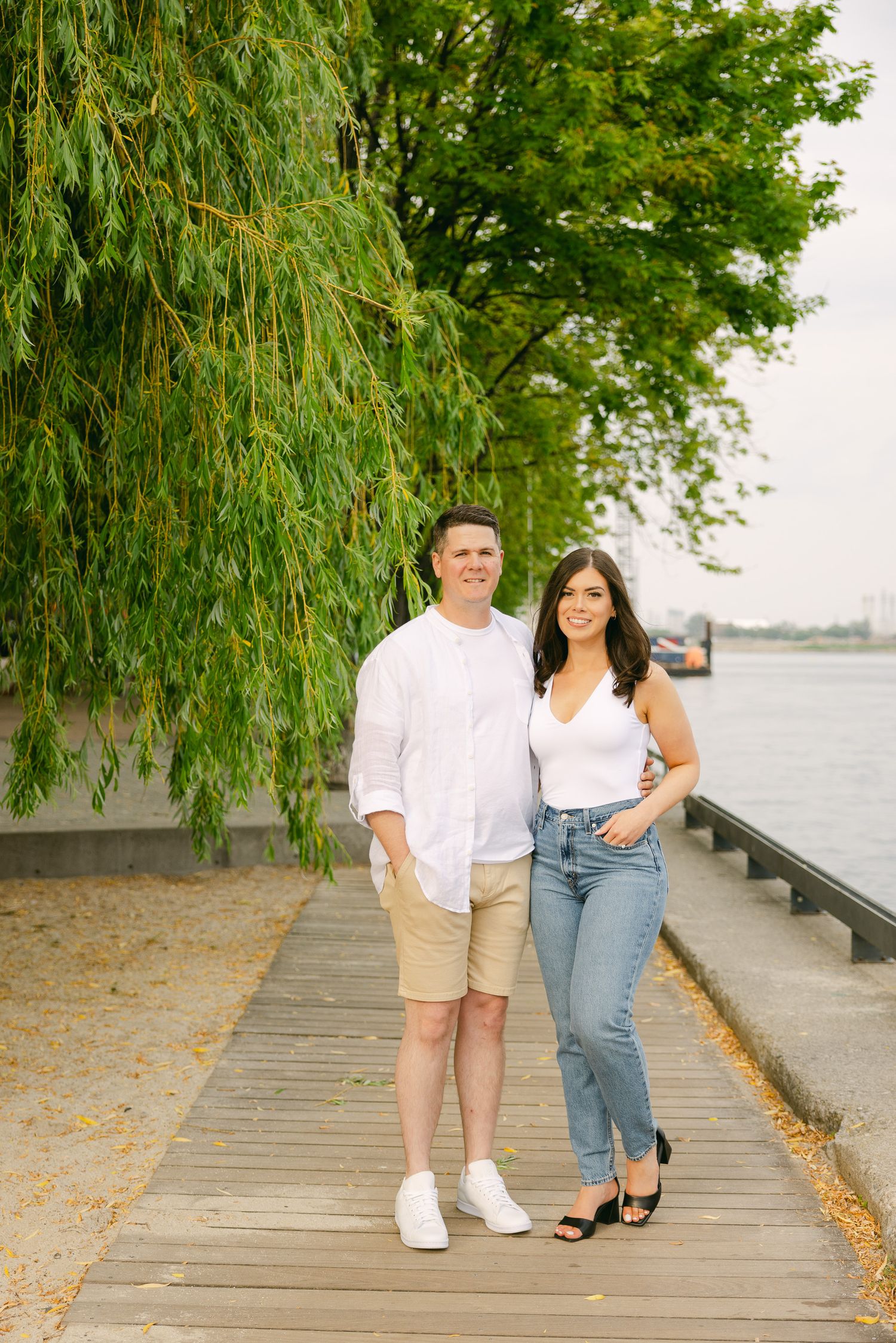 Stylish couple poses together on wooden boardwalk by water with willow tree in background.