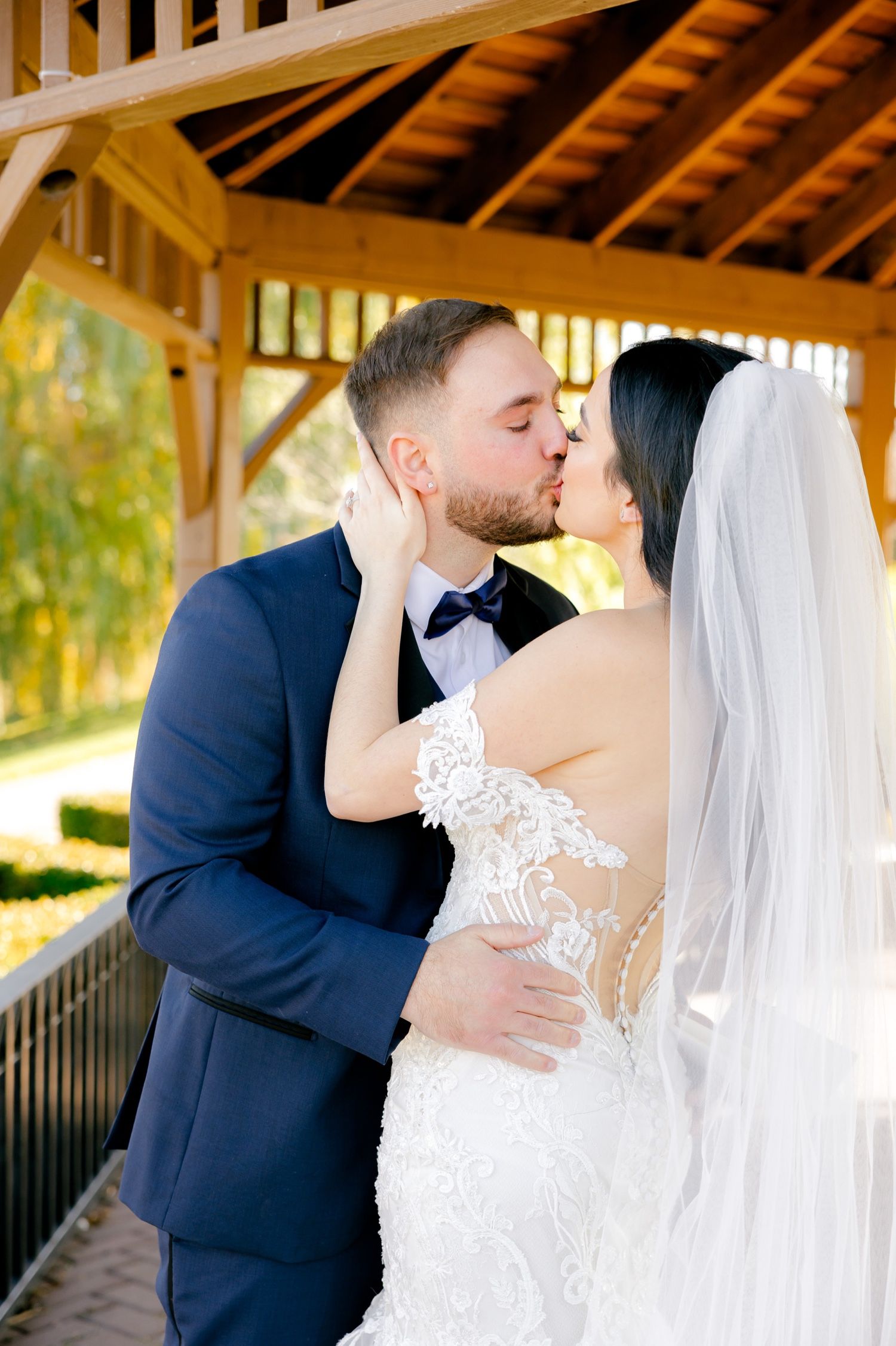Romantic wedding kiss under a wooden gazebo with sunlight streaming through.