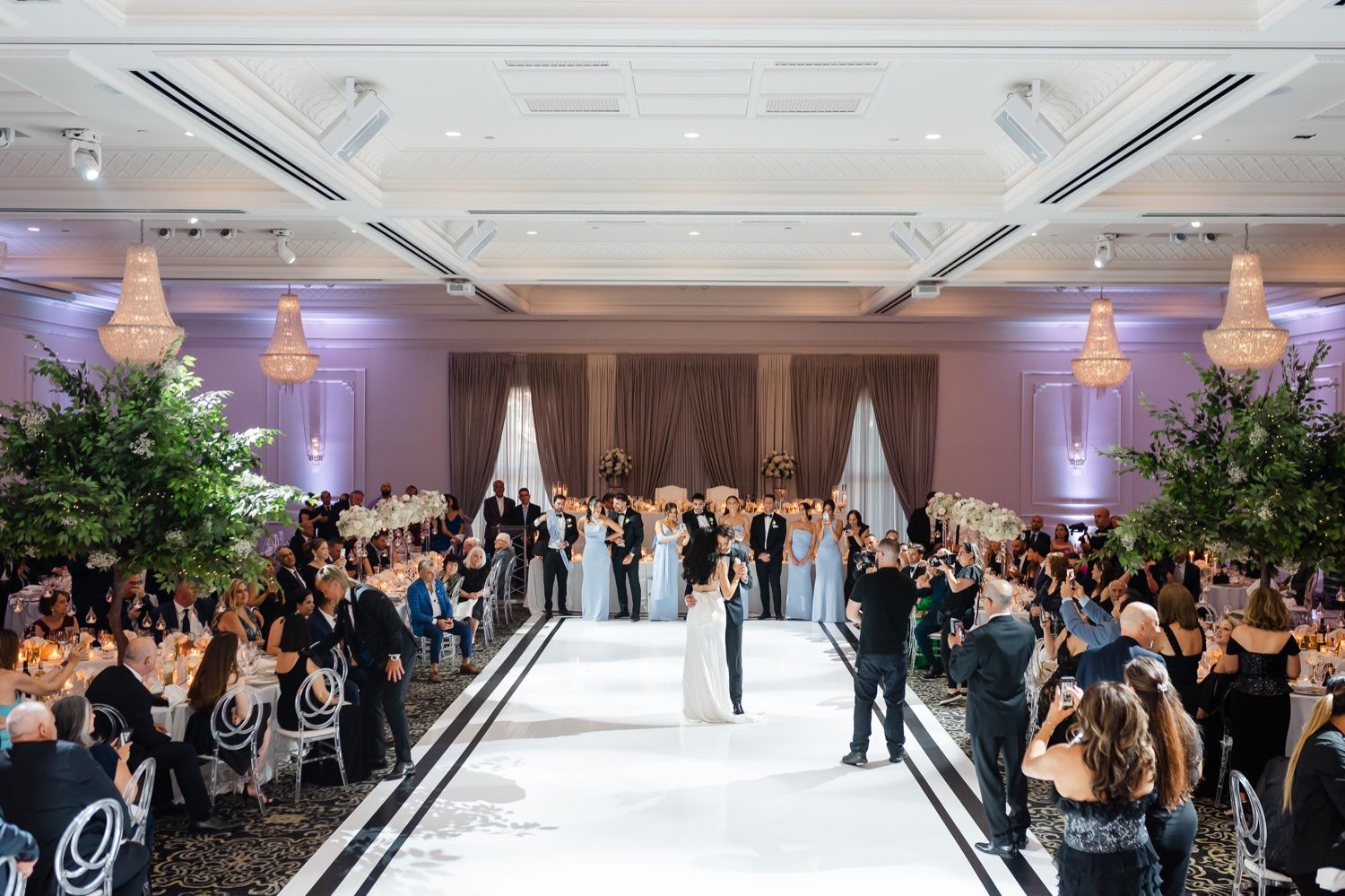 Guests cheer as couple makes their grand entrance into elegant ballroom with purple lighting.