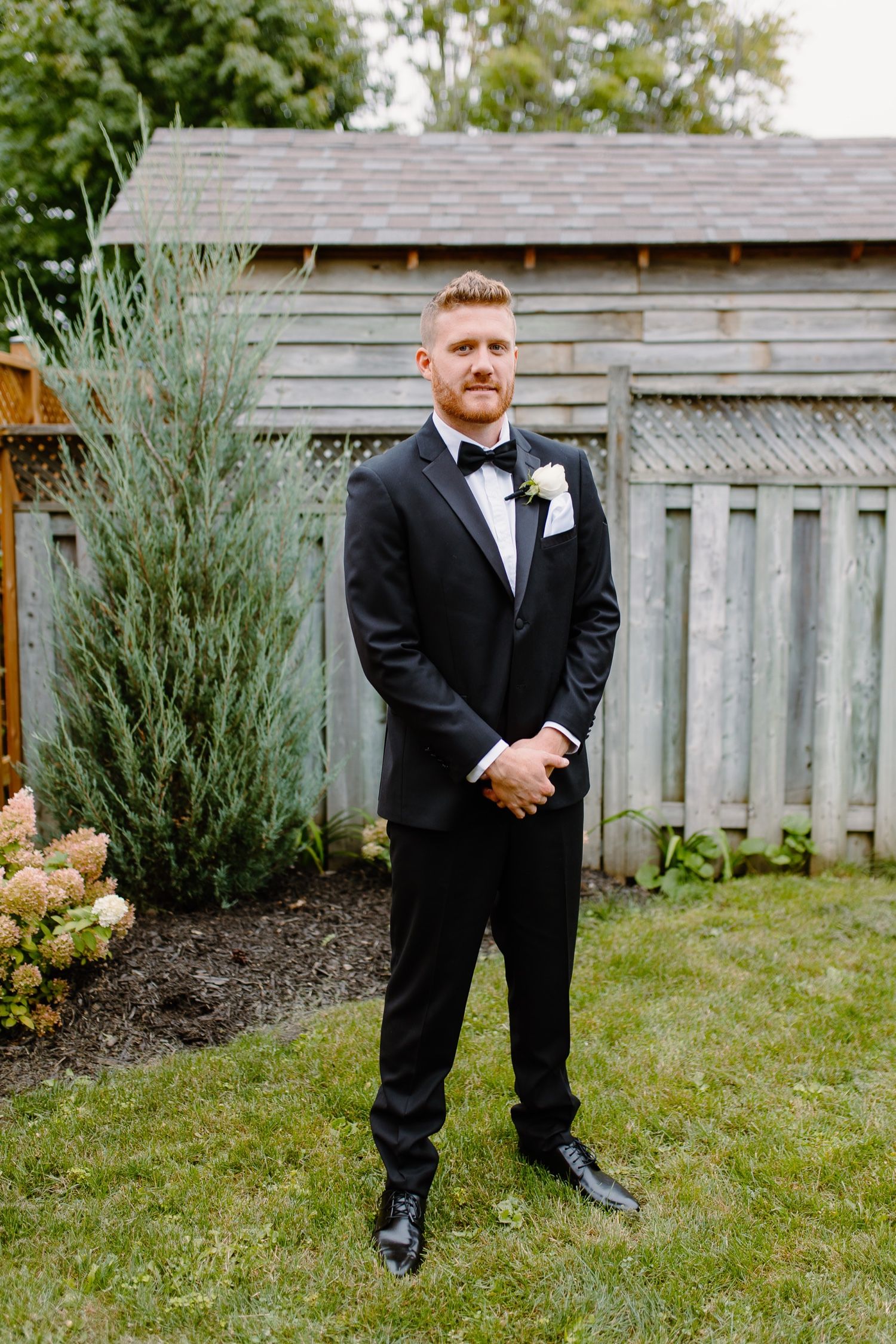 Groomsman in classic black tuxedo poses formally in garden setting with wooden fence backdrop.