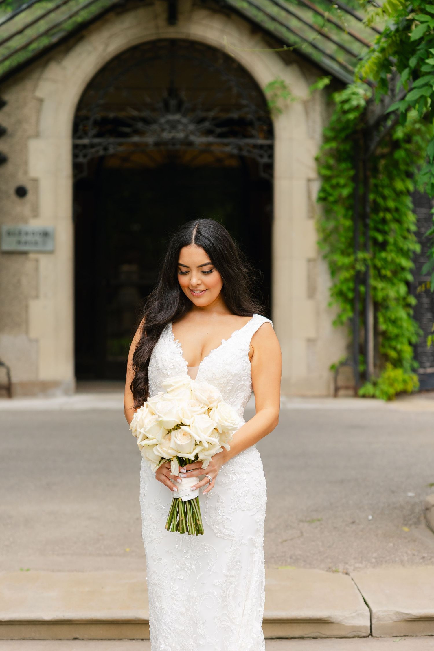 Close-up of bride holding white rose bouquet under stone archway surrounded by lush greenery.