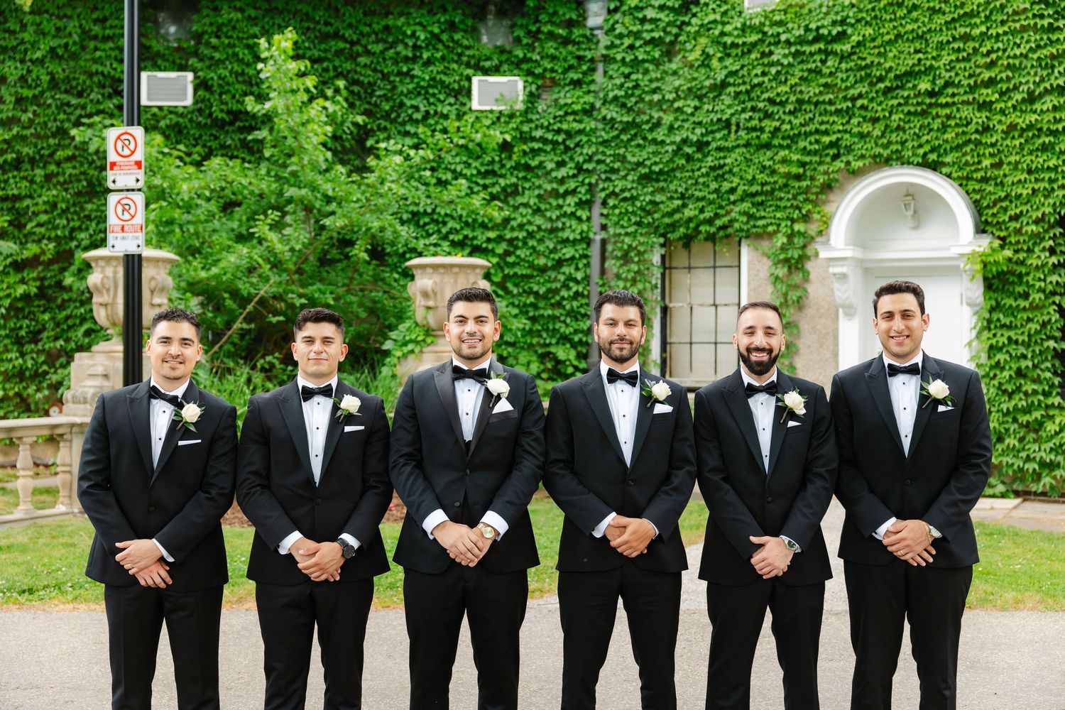 Wedding party in black tuxedos stands together against lush green ivy wall background.