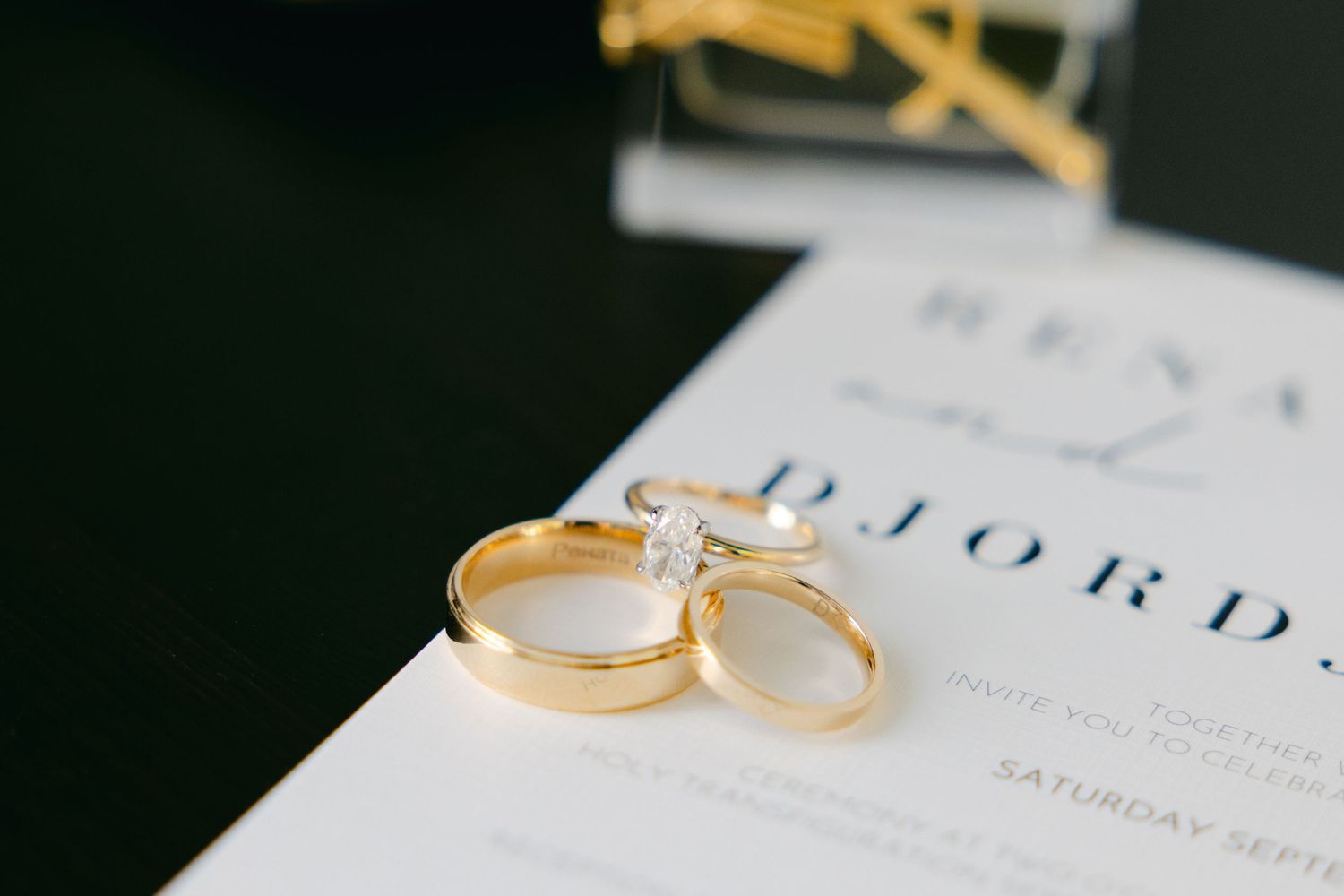 Gold wedding rings rest on a white wedding invitation against a dark background.