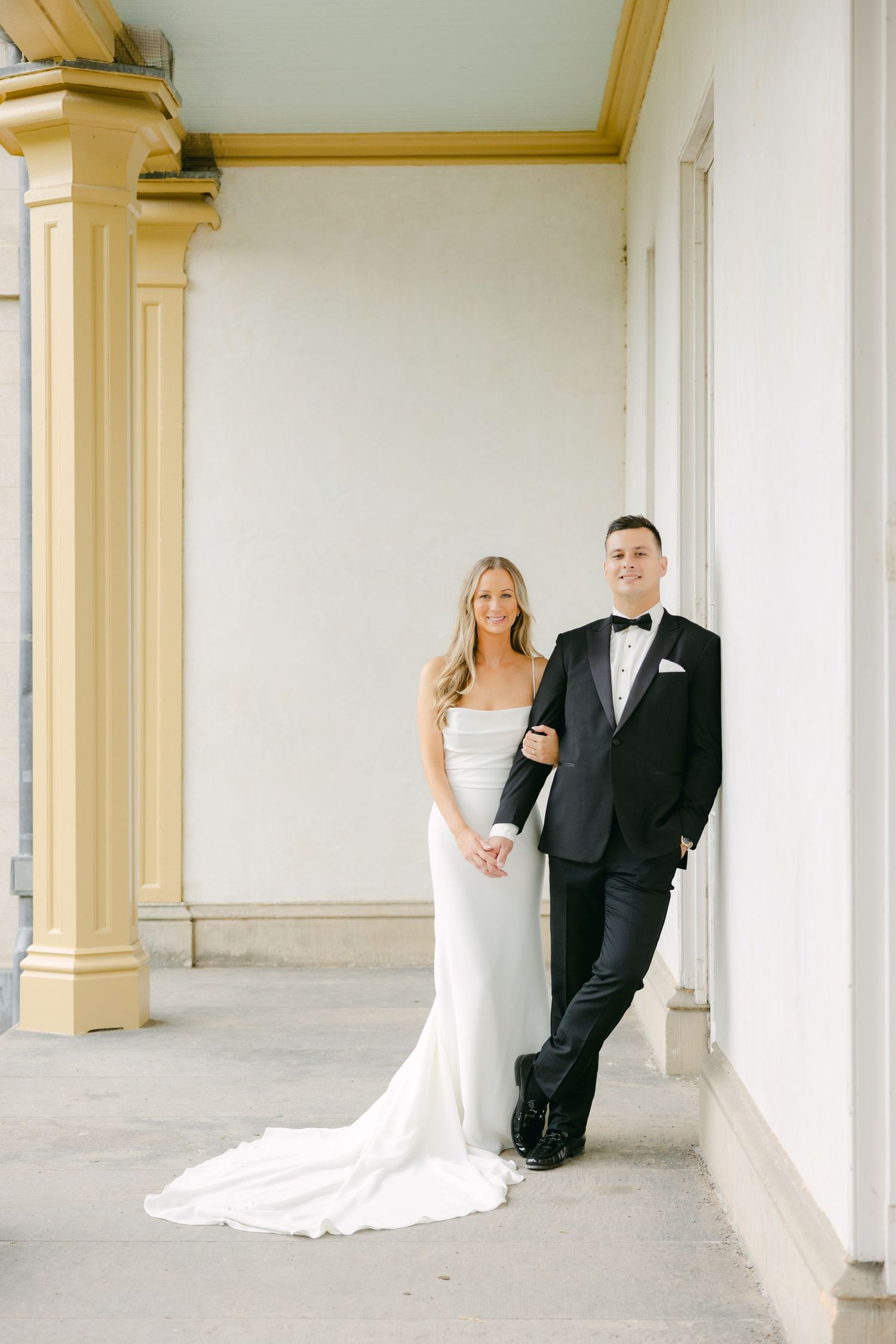 Elegant couple in wedding attire posing against white wall with golden architectural details.