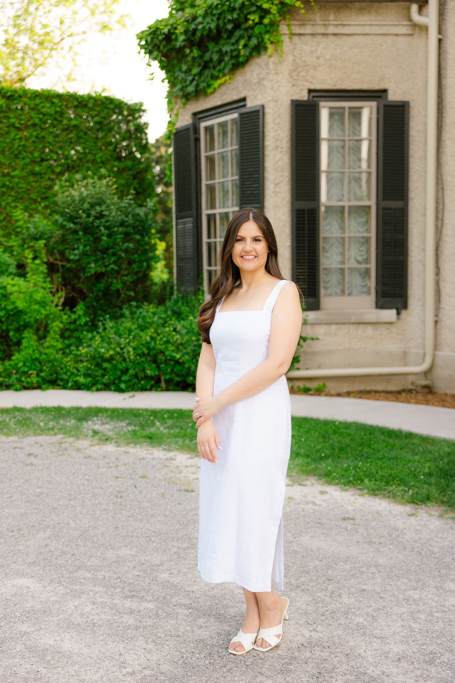 Woman in white midi dress stands in front of historic building with black shutters and ivy.